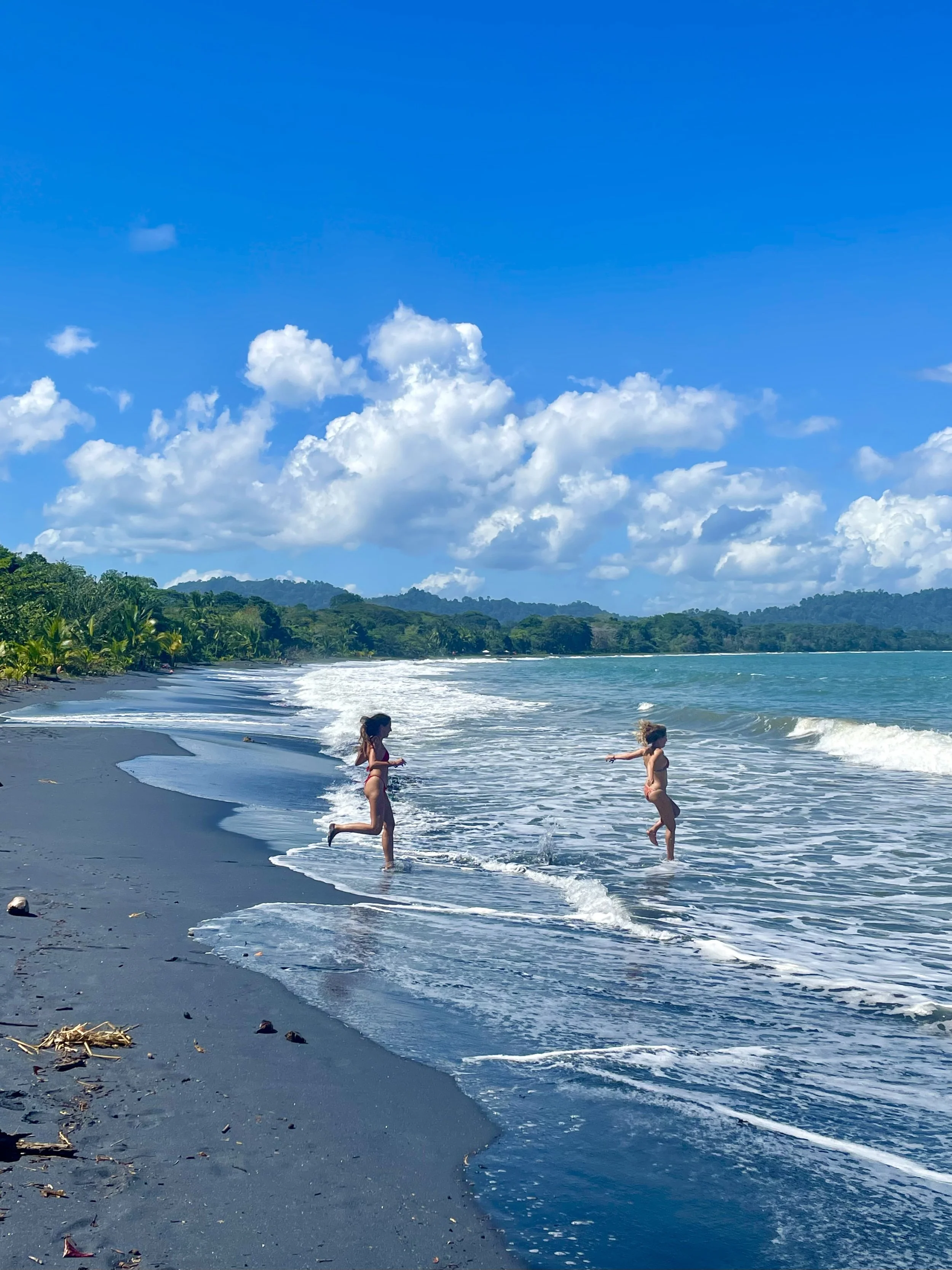 Two children playing in the ocean waves on a sandy beach with a blue sky and fluffy clouds in the background.