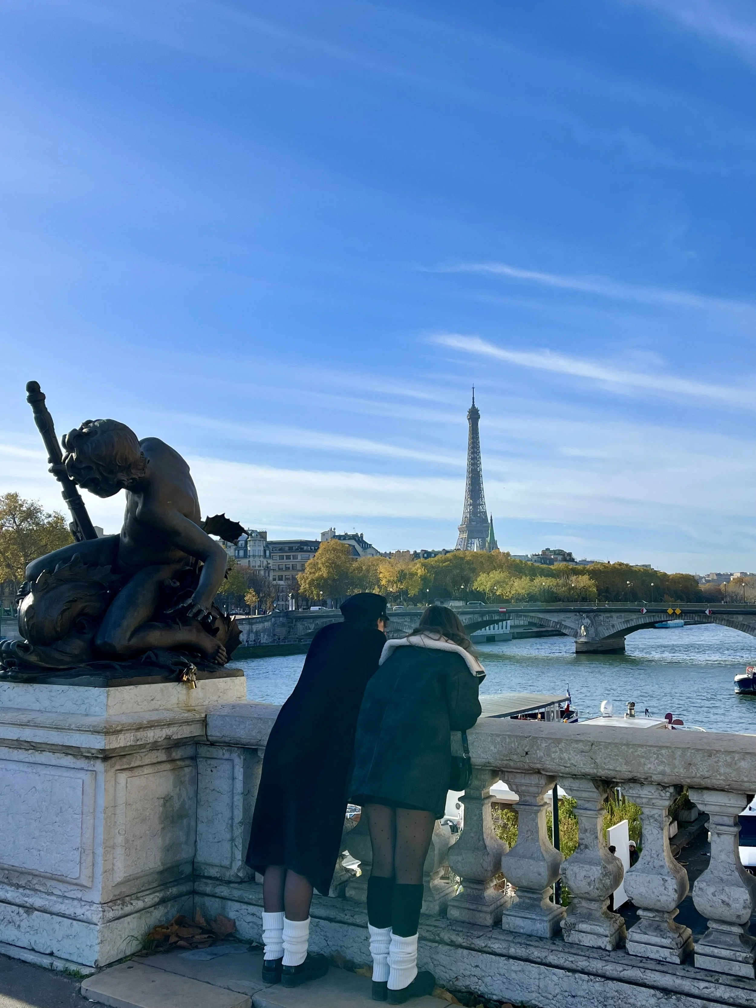 Two people leaning against a stone railing on a bridge overlooking the Seine River, with a statue in the foreground and the Eiffel Tower in the background under a clear blue sky.
