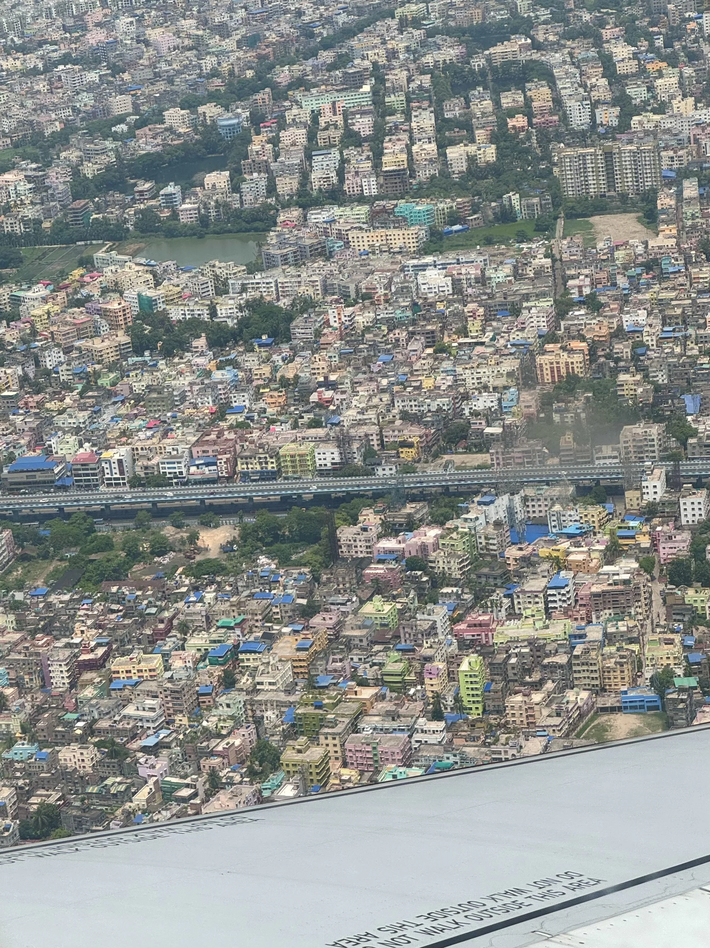 Aerial view of a densely populated city with colorful buildings, roads, and greenery, partially obscured by an airplane wing.
