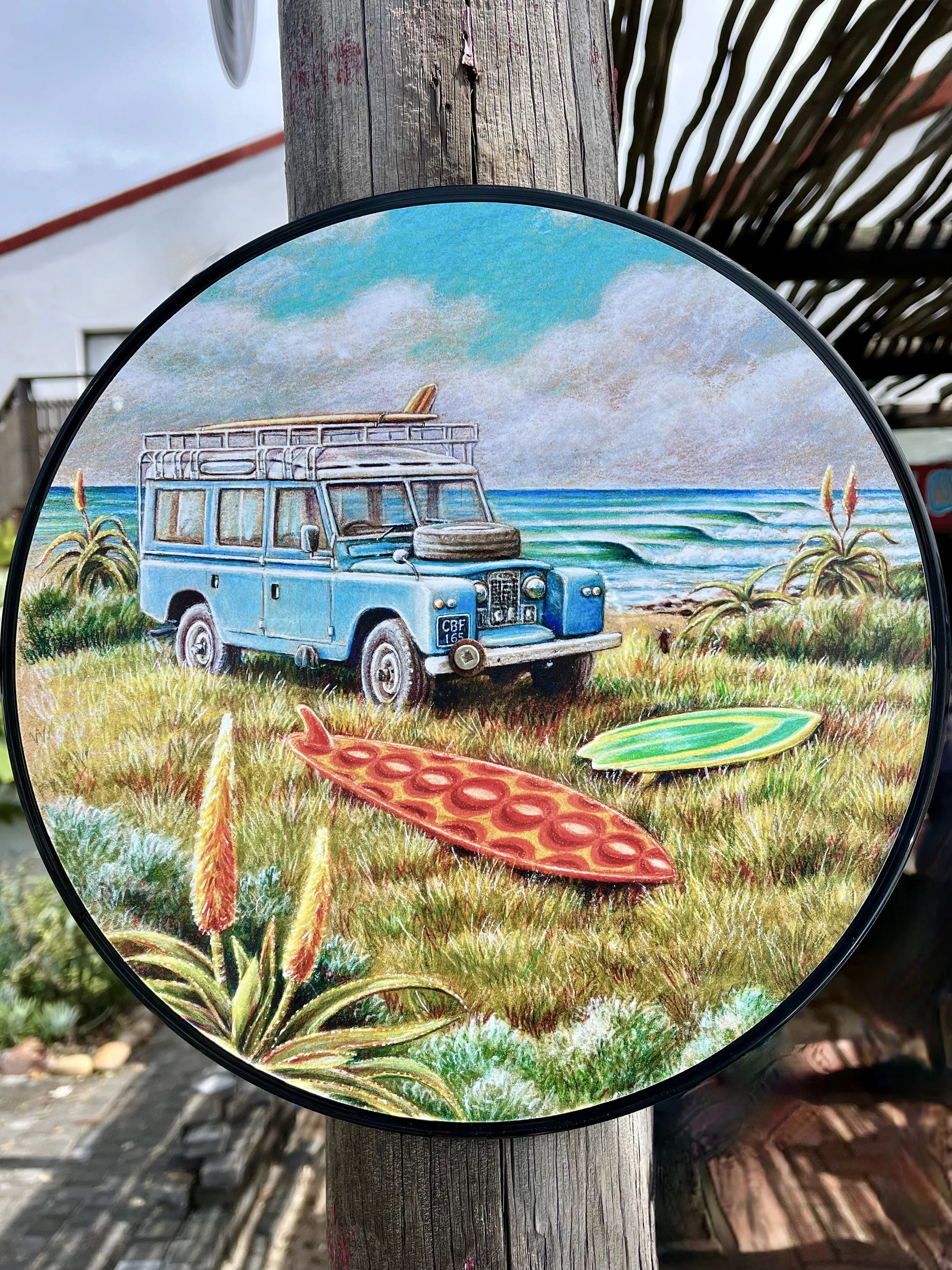 Painting featuring a vintage blue Land Rover parked near the ocean with surfboards on the grass and a beach in the background.