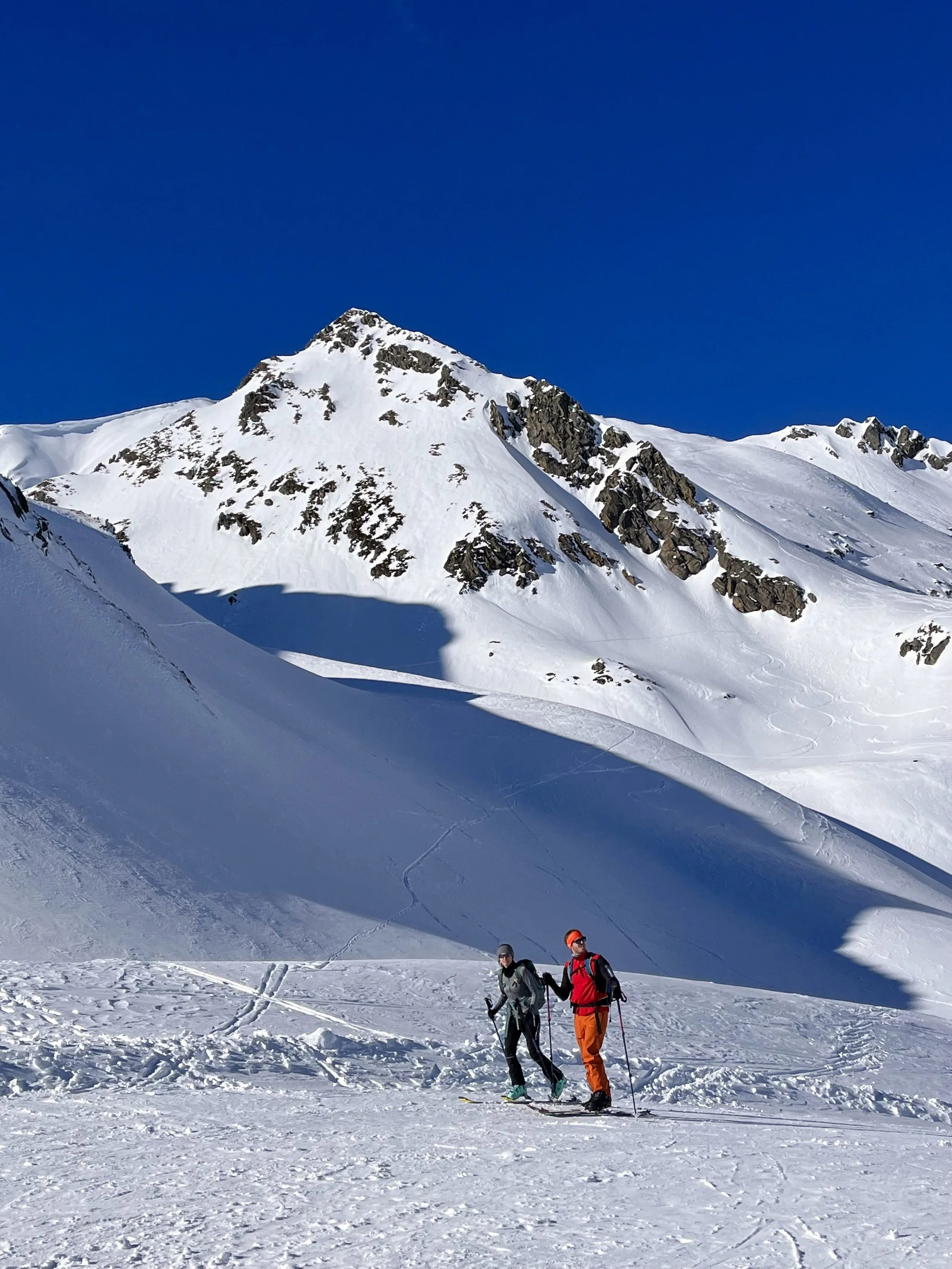 Two skiers on a snowy mountain with a clear blue sky and rocky peaks in the background.