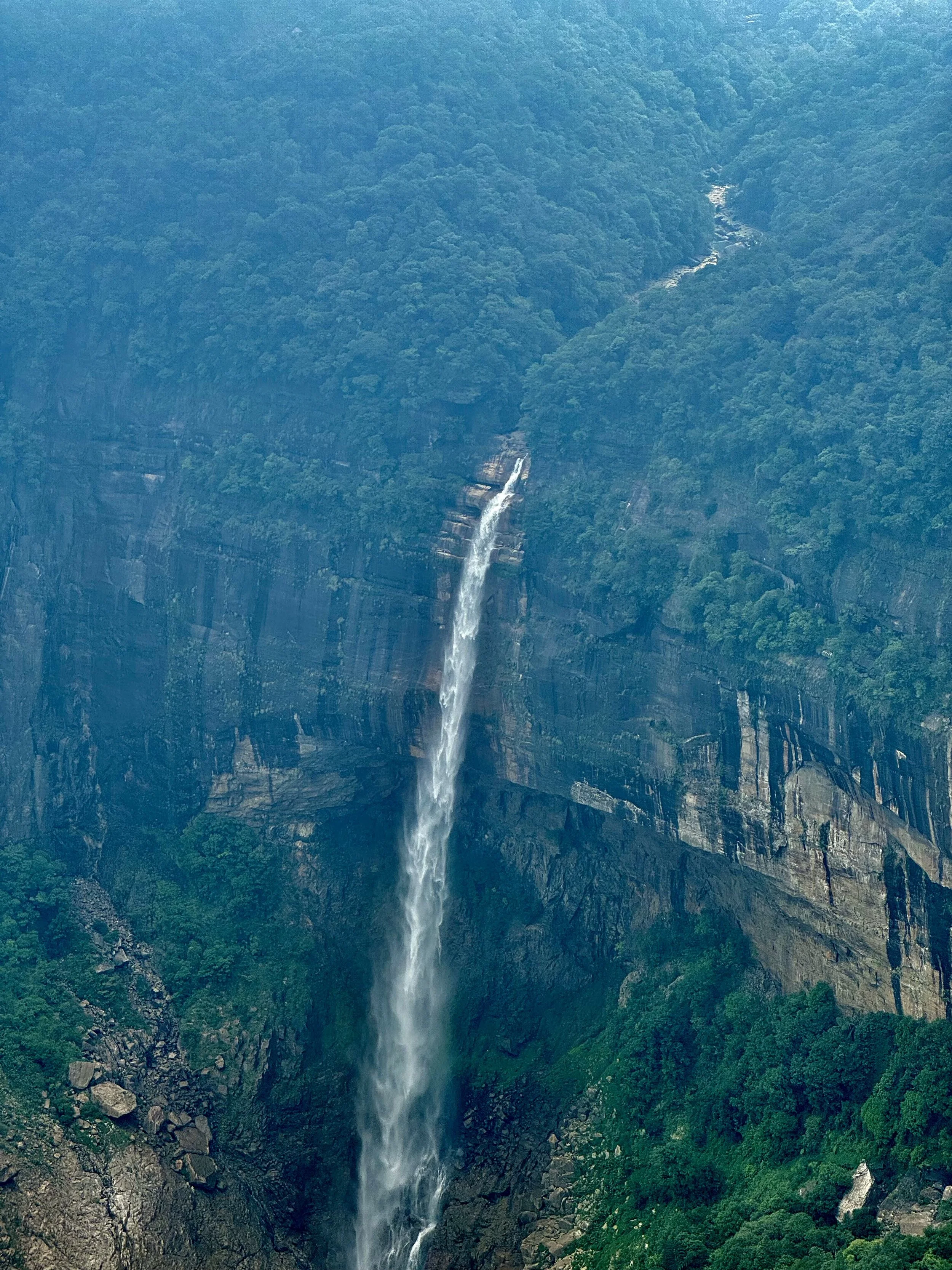 Tall waterfall cascading down a lush green cliff into a rocky forested valley.