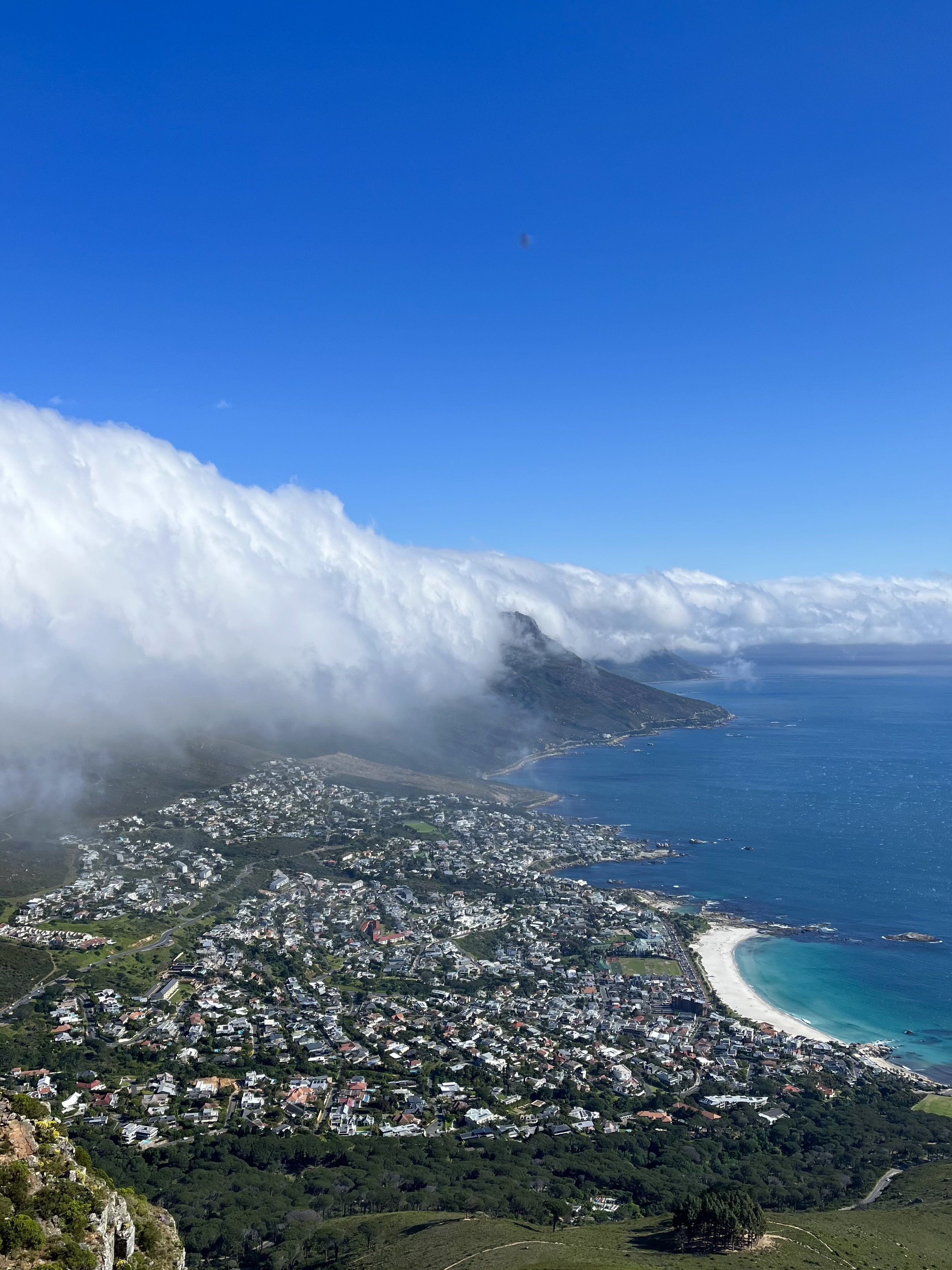 Aerial view of a coastal city with clouds, green hills, and a beach along a blue ocean.