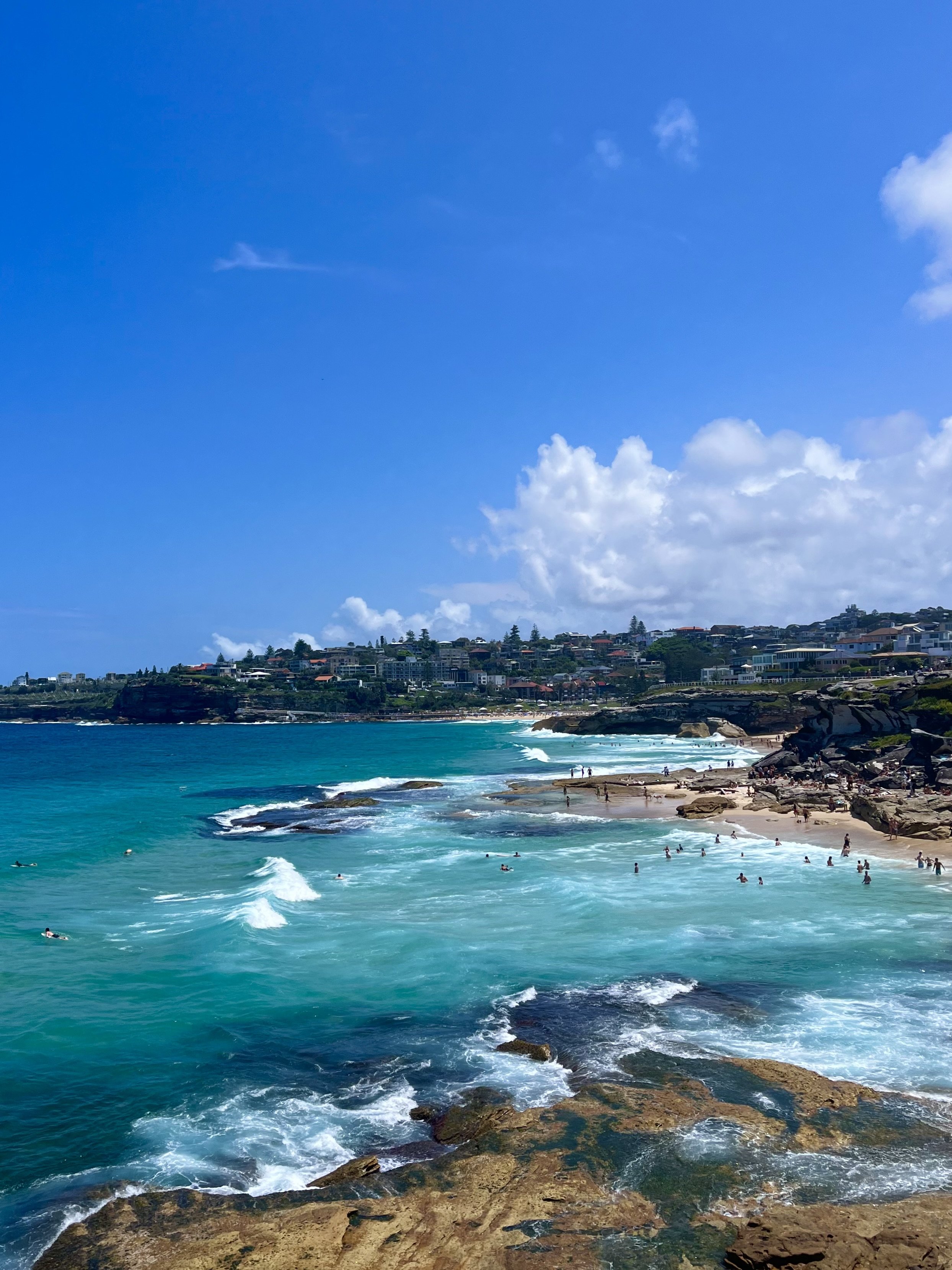 A scenic coastal view of a beach with turquoise waters, waves crashing on rocks, and people swimming and enjoying the sandy shoreline. The sky is clear and blue with a few clouds, and a residential area with houses can be seen in the background on a 