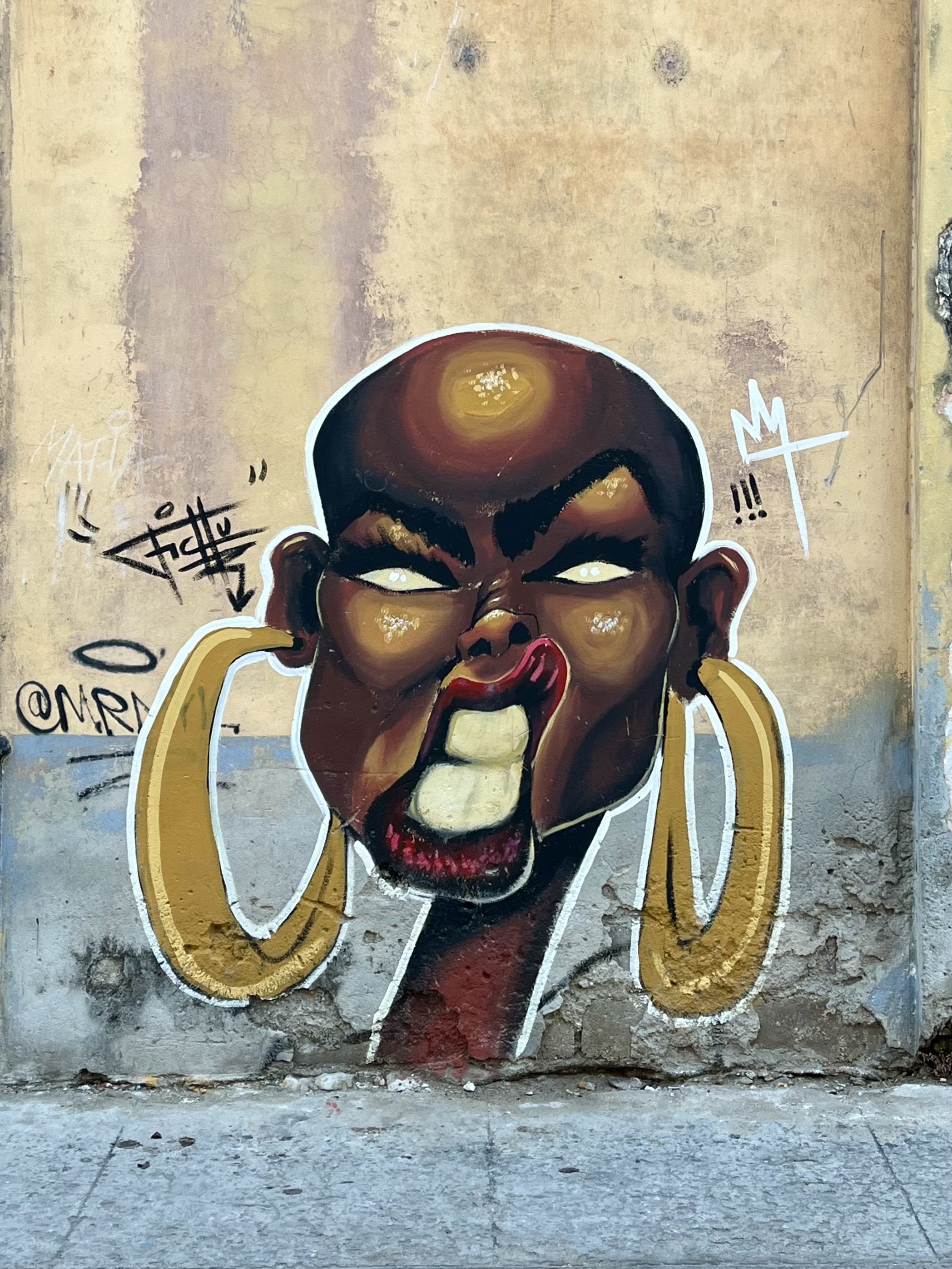 Street art graffiti of a stylized face with large hoop earrings and an exaggerated expression on an old wall.