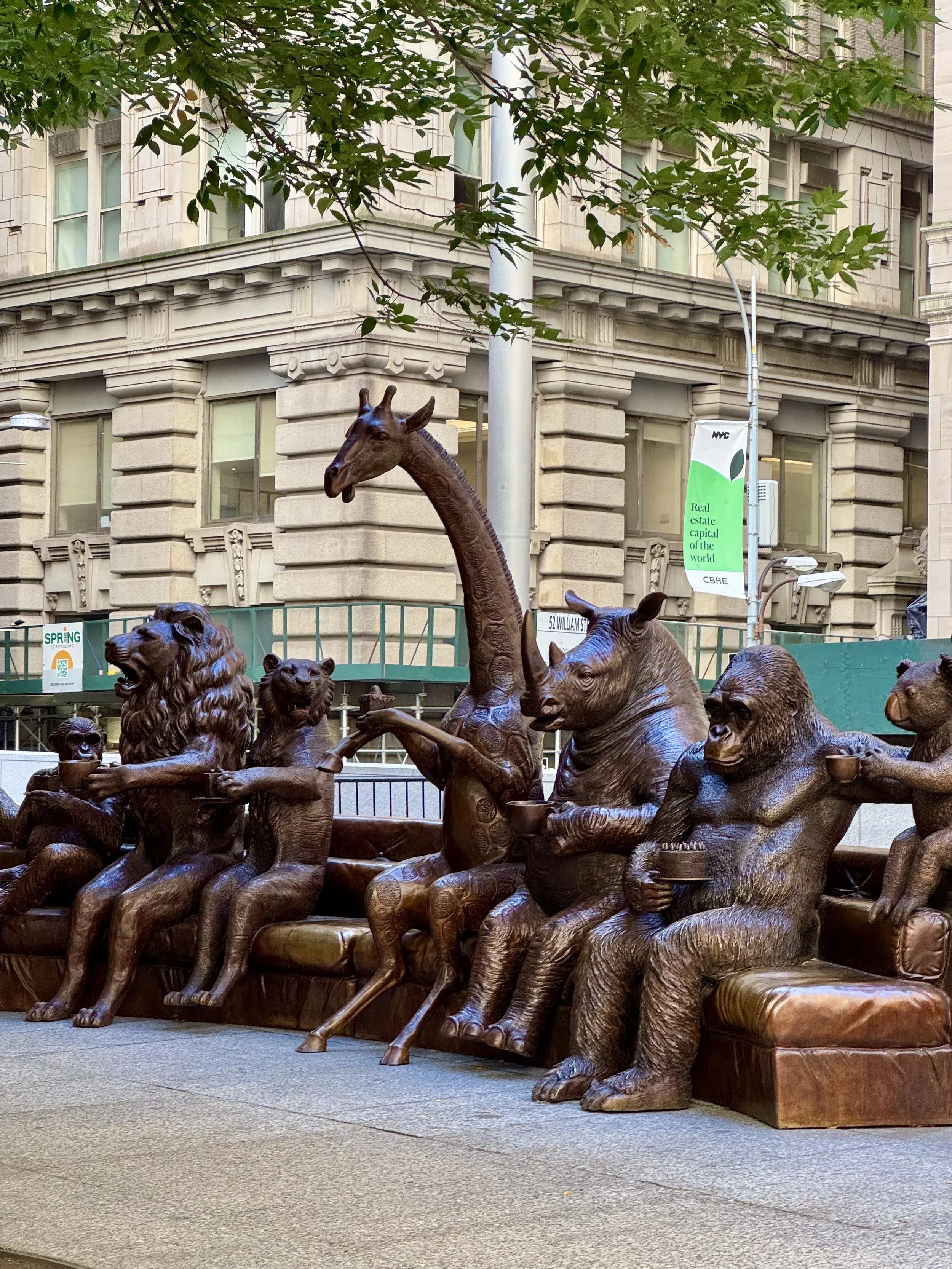 Bronze animal sculptures sitting on a bench in a city setting, including a gorilla, giraffe, and bear holding objects like cups.