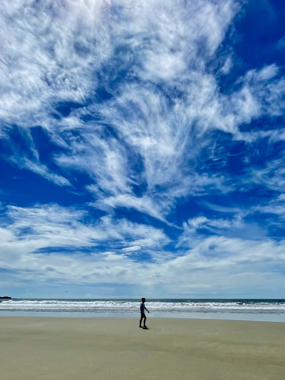 Person walking on a sandy beach with ocean waves, under a blue sky with wispy clouds.