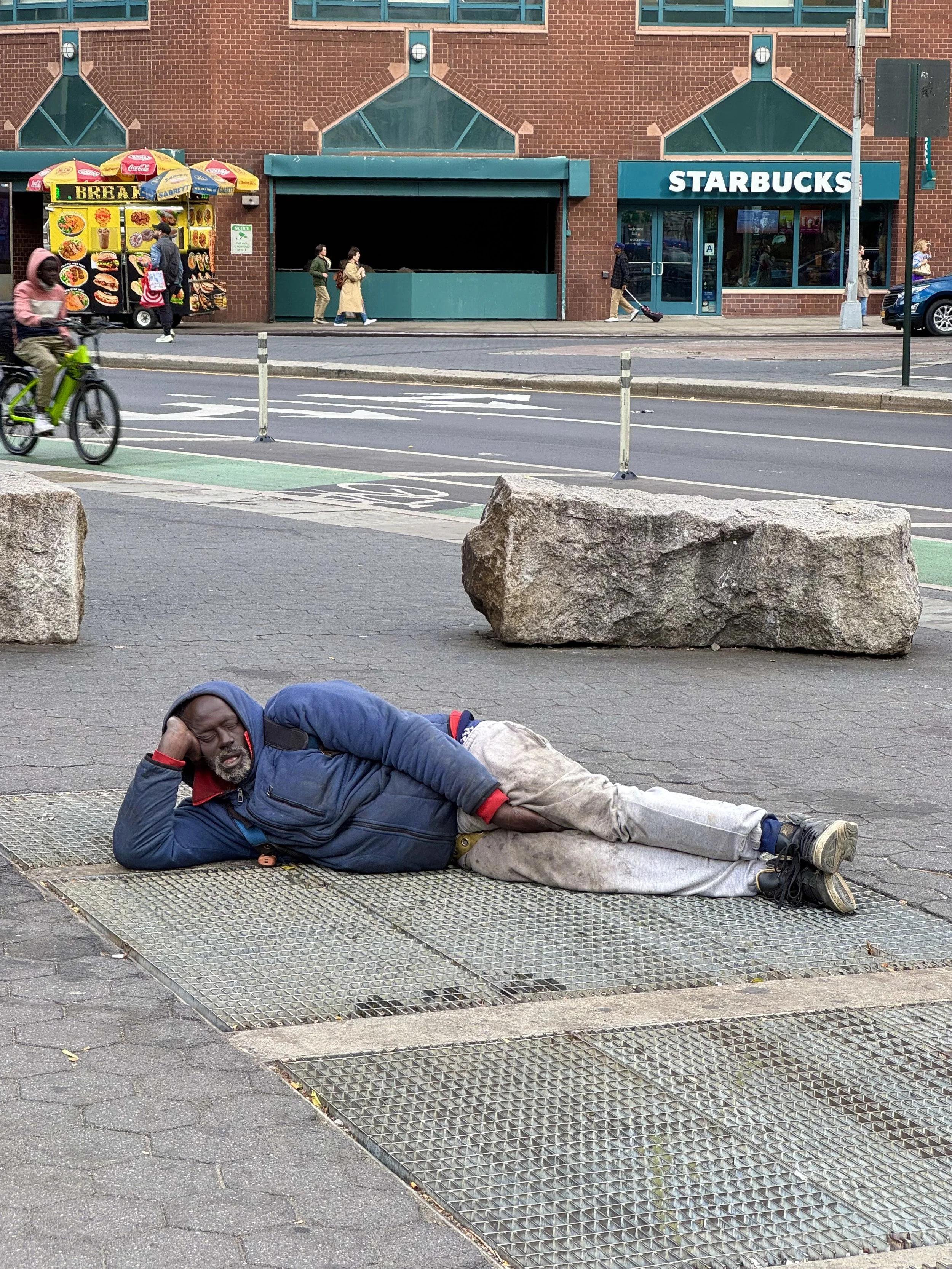 Person in a blue jacket lying on a sidewalk in front of a Starbucks, with a bike lane and food truck nearby.