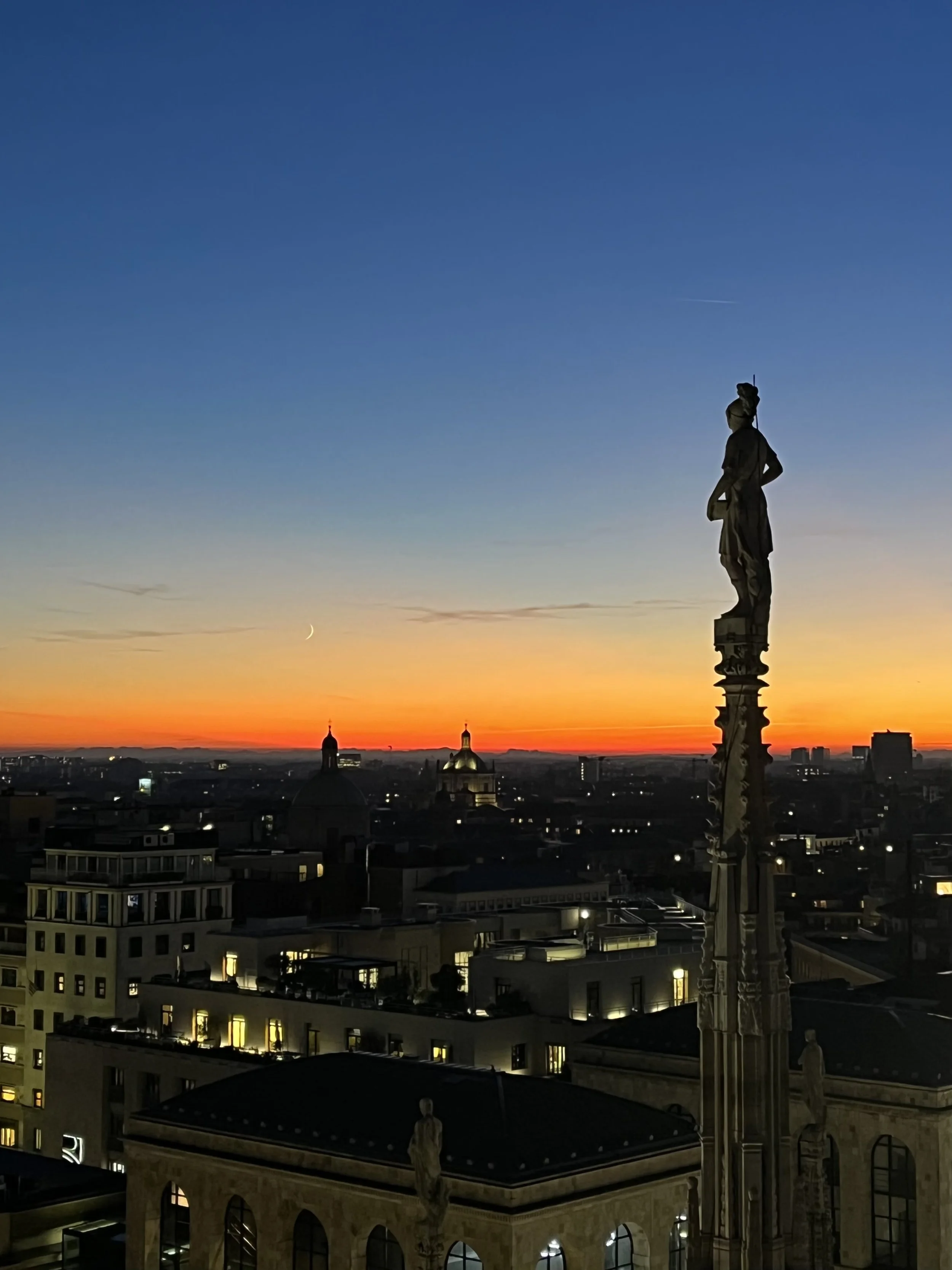 Rooftop view at sunset with skyline silhouettes and a prominent statue against a colorful sky.