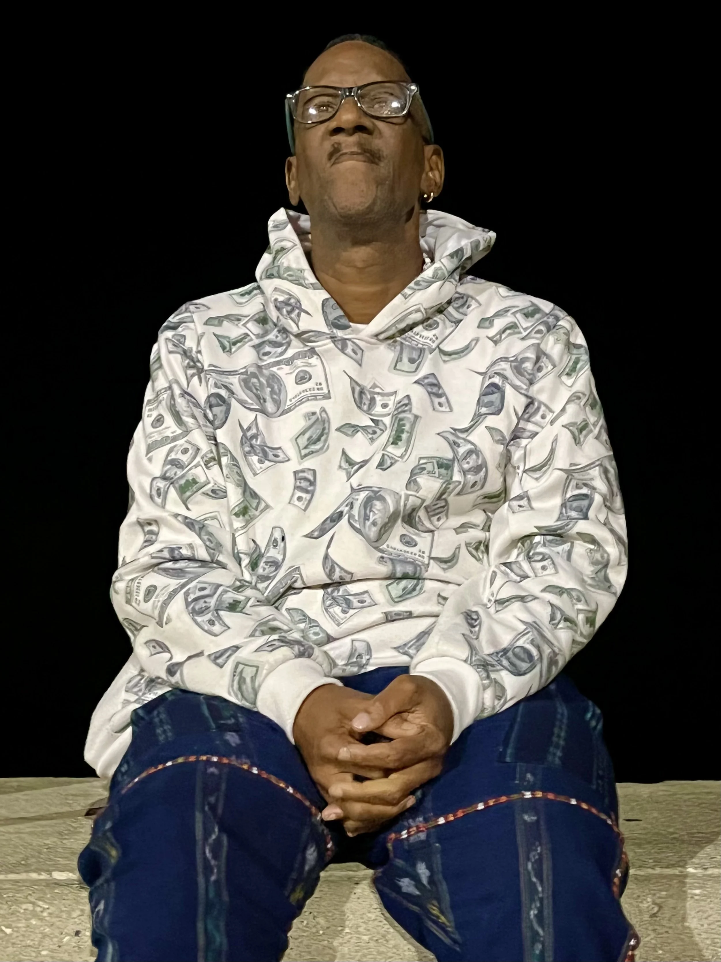 Person wearing glasses and a hoodie with dollar bill print, sitting against a dark background.