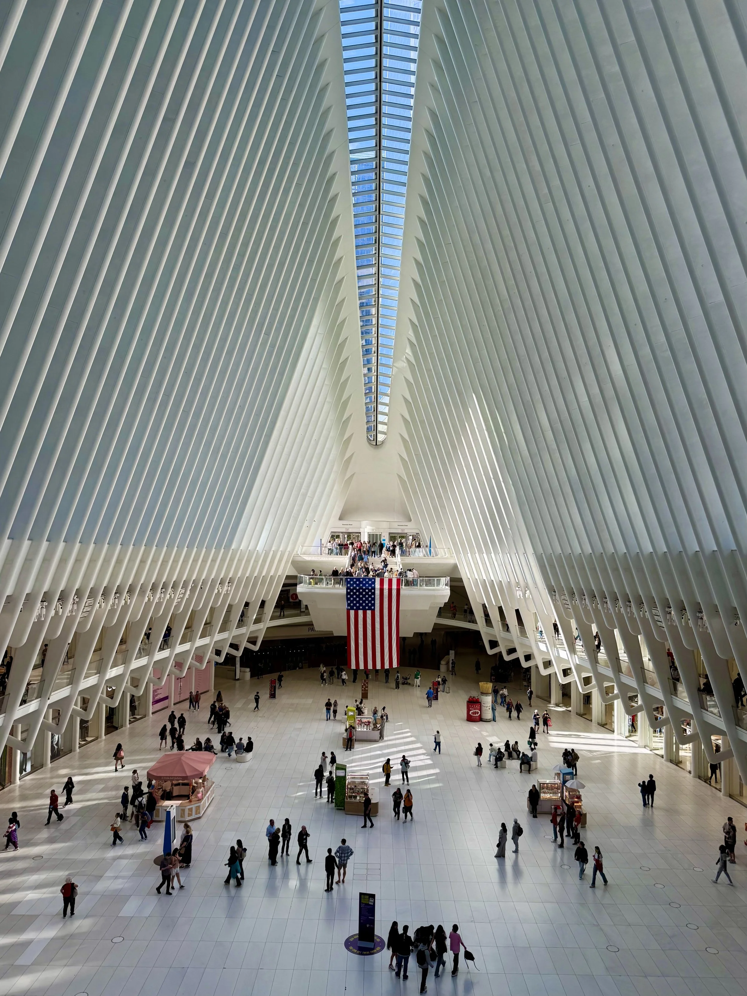 Interior of the Oculus transportation hub at the World Trade Center in New York City, featuring high white ribbed arches, a large American flag, and people walking on the floor below.