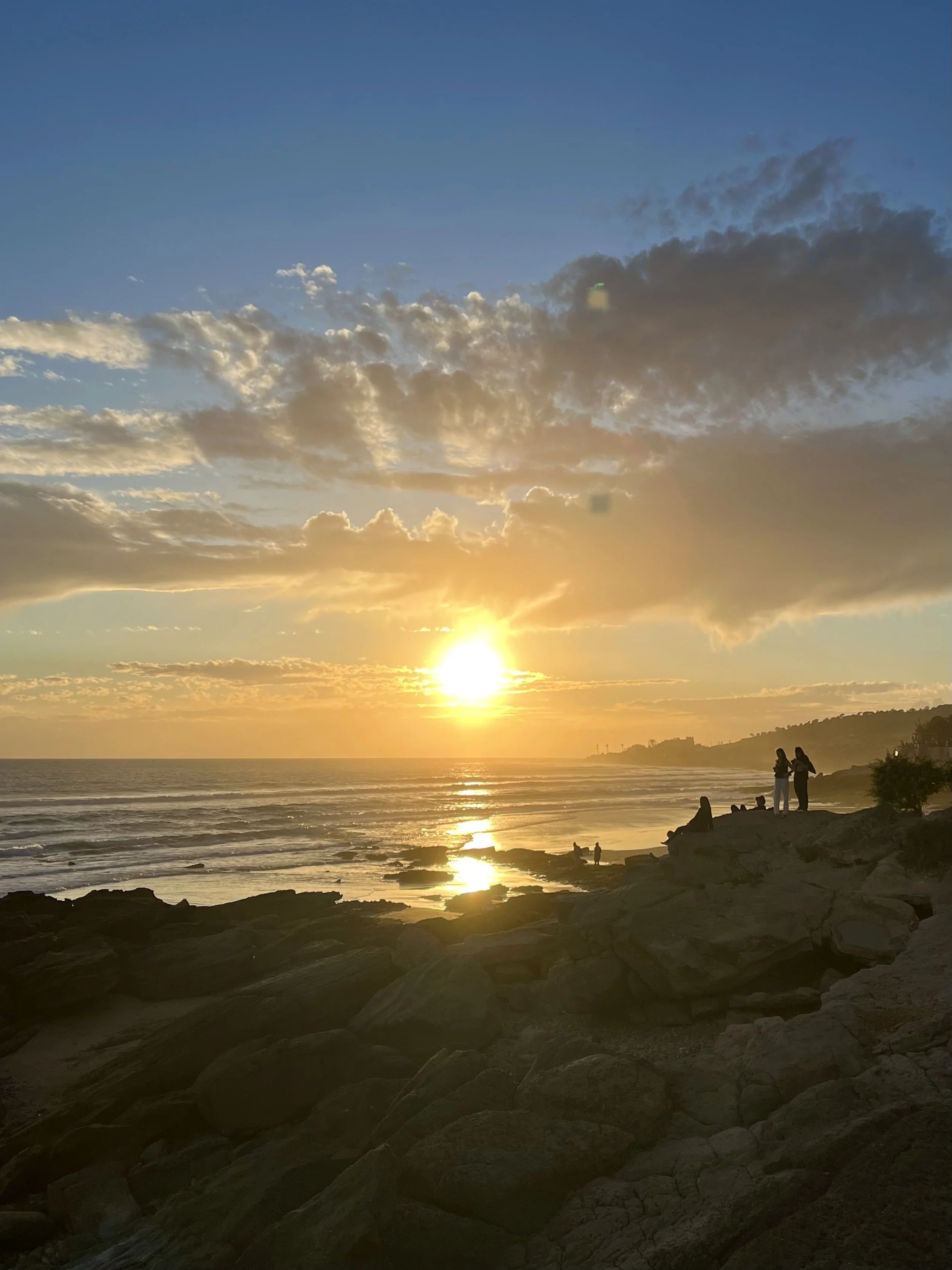 Sunset over rocky beach with silhouetted people, ocean waves, and partly cloudy sky.