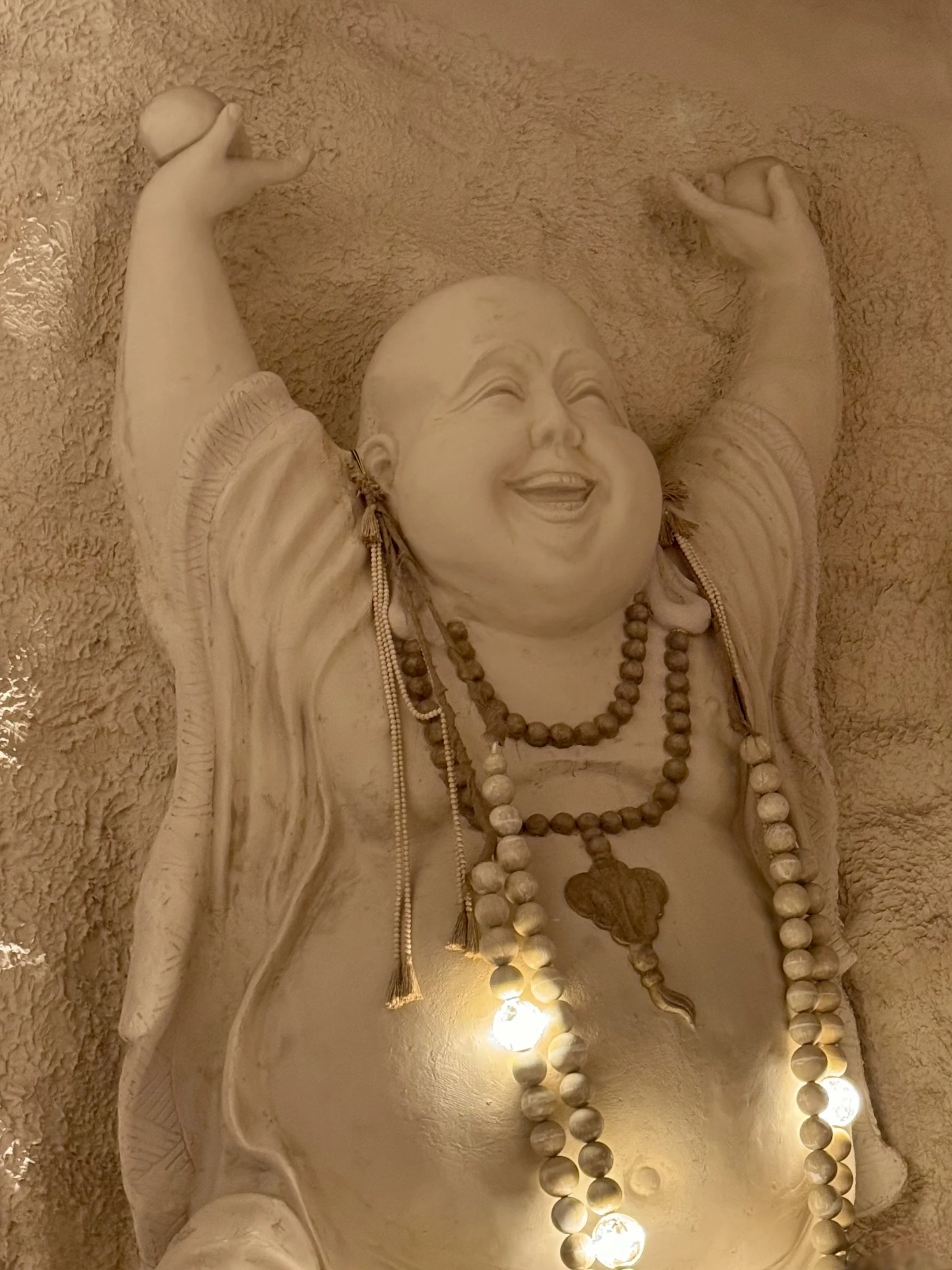 Smiling statue of Budai with raised arms, wearing beaded necklaces, against a textured wall.