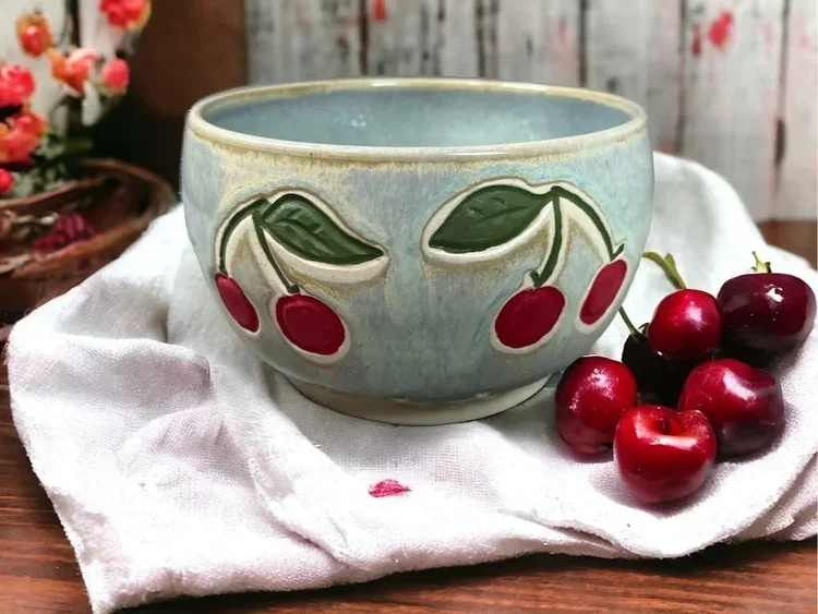 A ceramic bowl with a Cherry design sits on a white cloth, with fresh cherries placed beside it, on a wooden table.