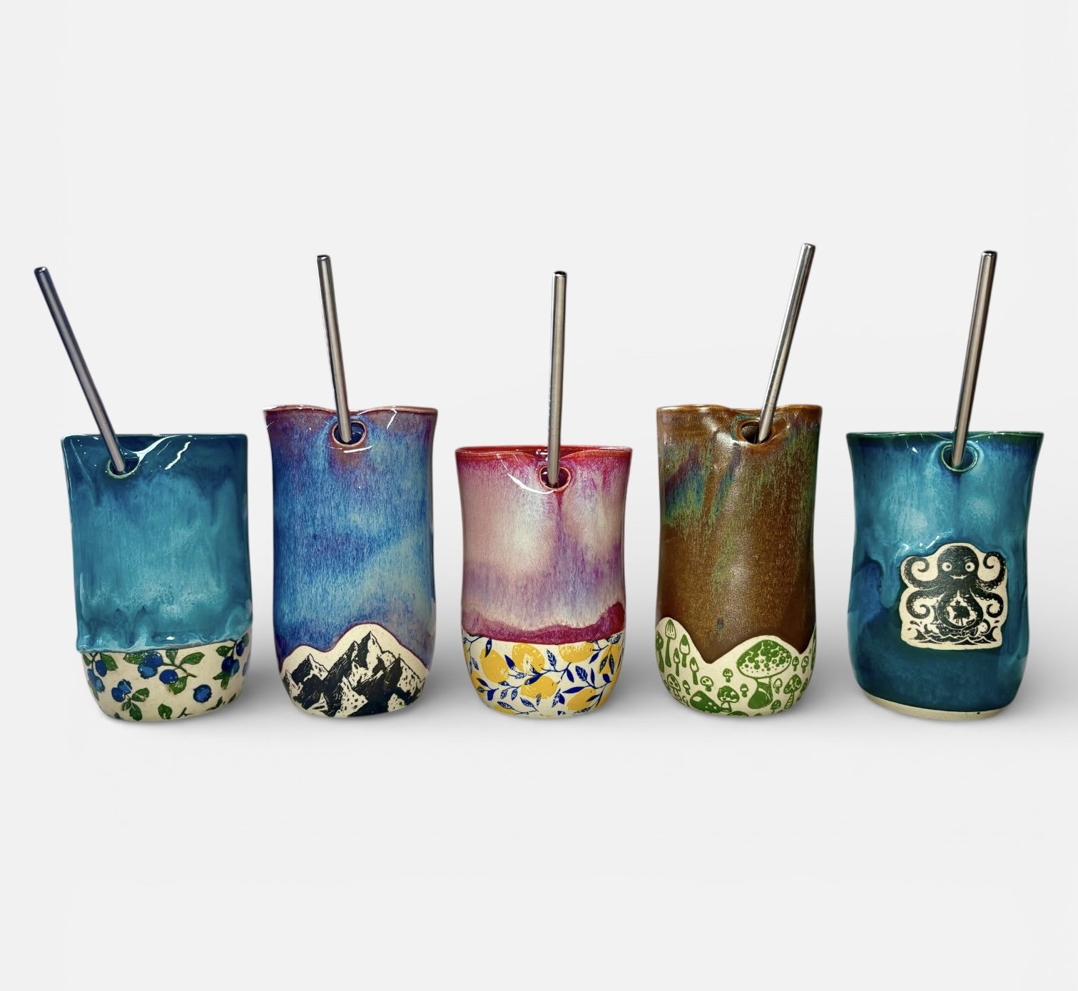Five colorful ceramic cups with metal straws, each featuring different artistic designs and patterns, arranged in a row against a white background.