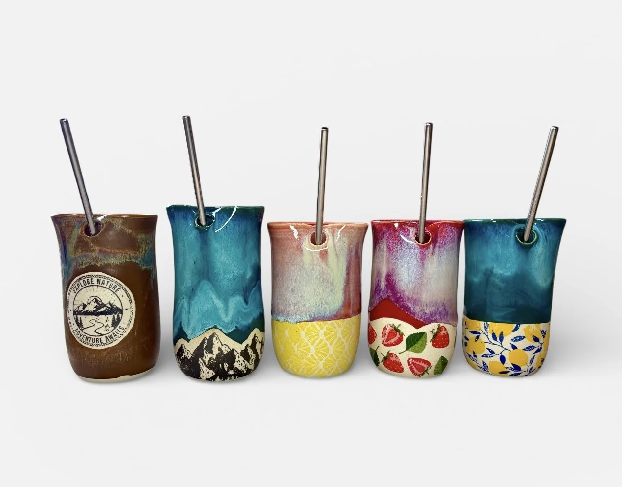 Five ceramic tumblers with colorful, artistic designs, each with a metal straw, lined up against a plain white background.