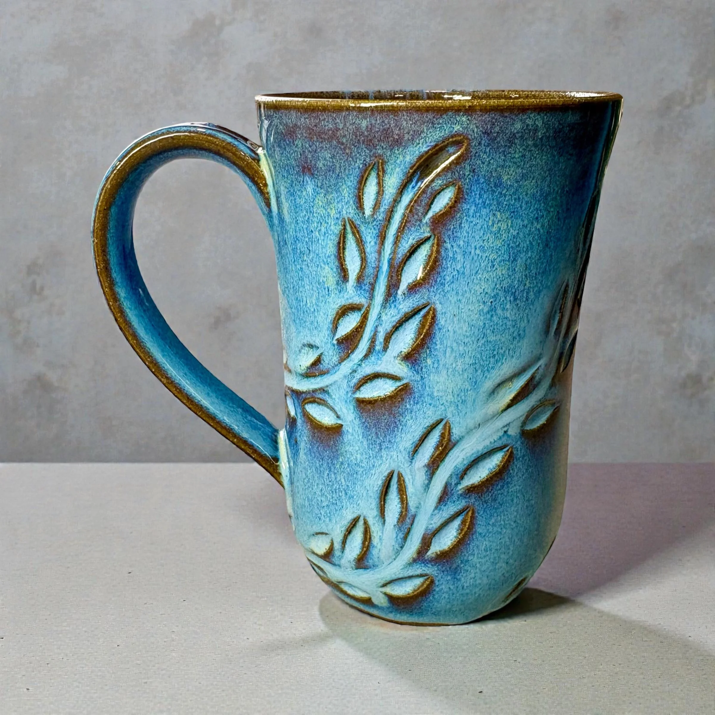 Ceramic mug with a blue glaze and carved leaf pattern design, sitting on a plain surface.
