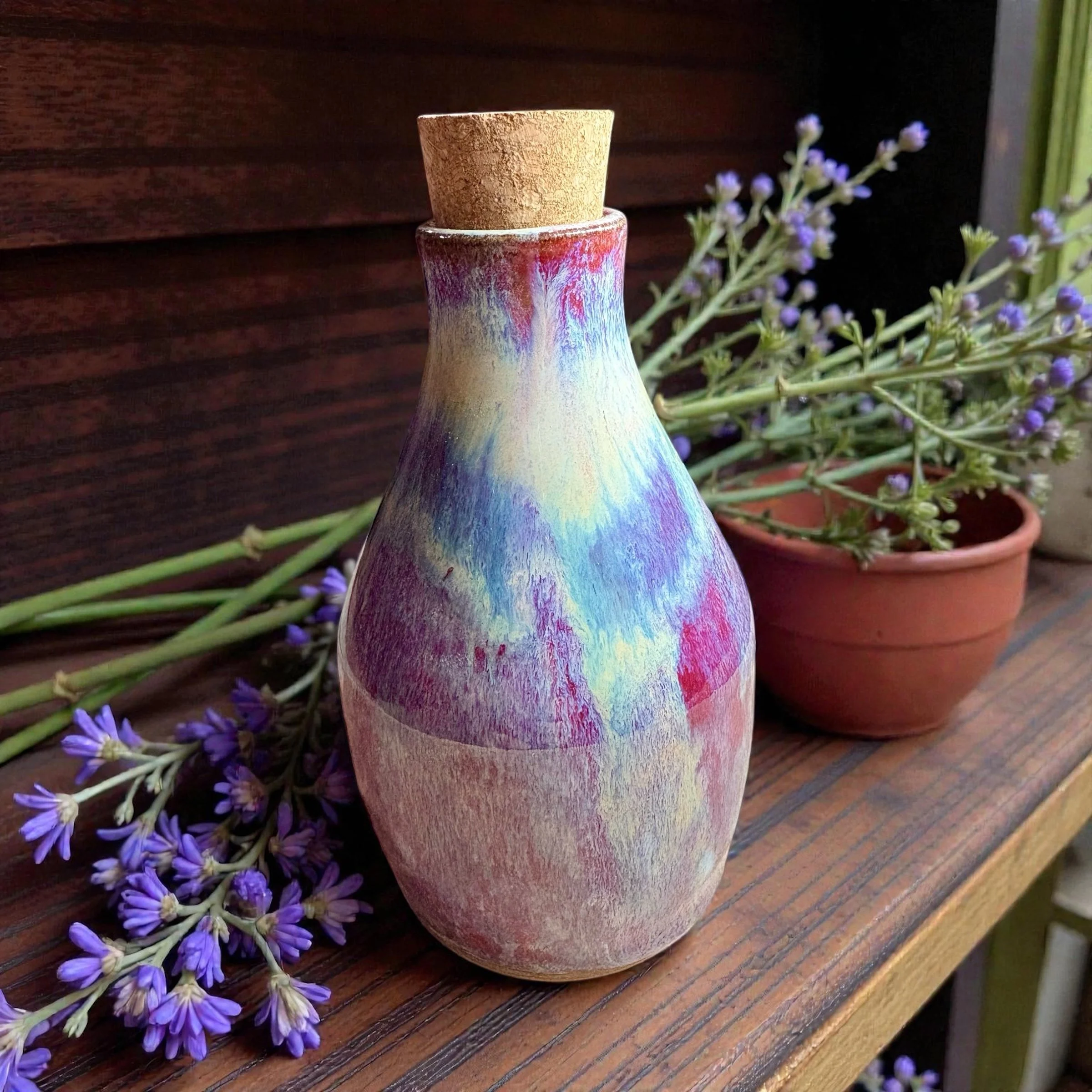 Colorful ceramic vase with a cork lid, placed on a wooden shelf next to potted purple flowering plant.