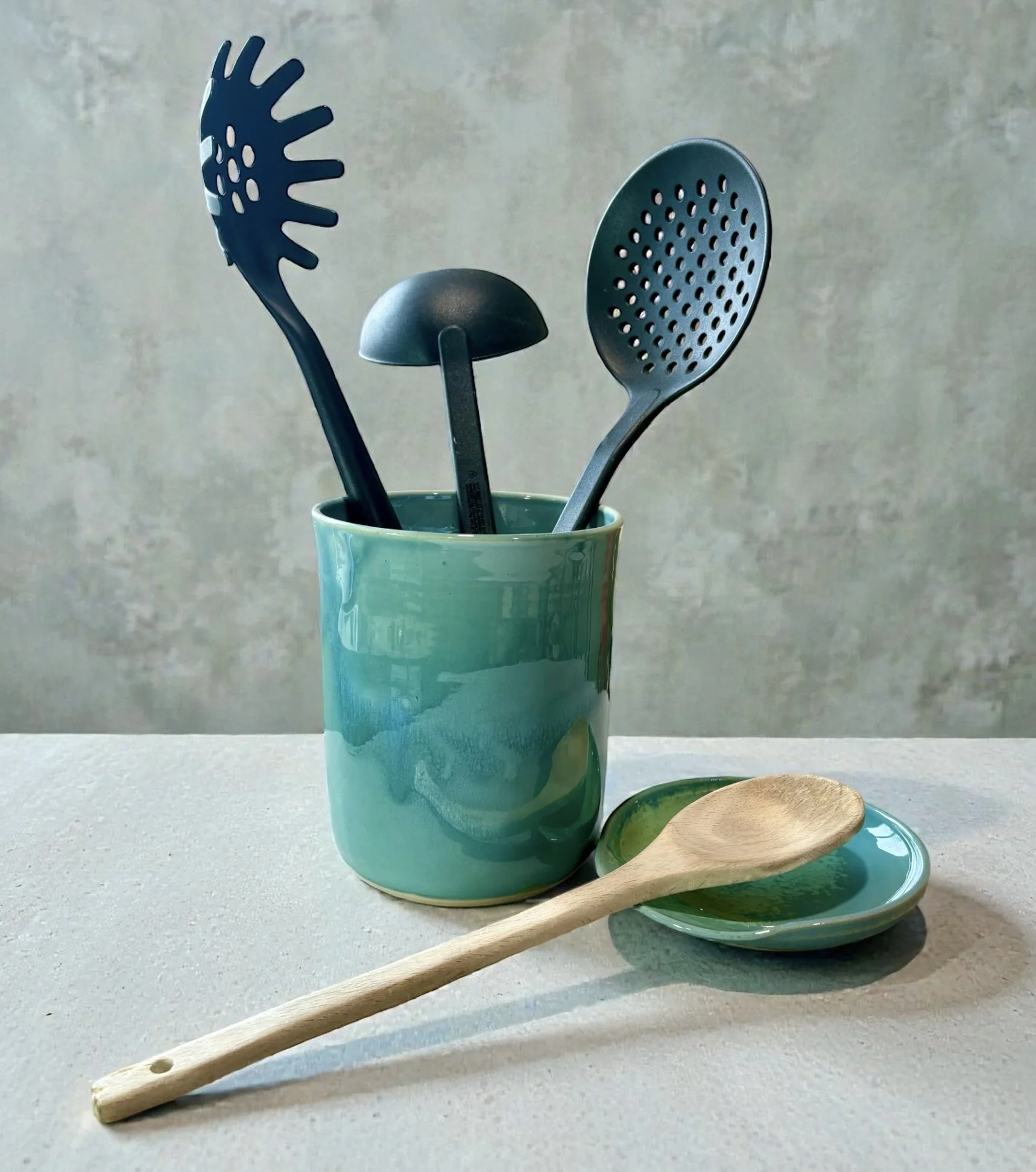 A turquoise ceramic cup holding three black kitchen utensils: a slotted spatula, a ladle, and a slotted spoon. In front of the cup are a small turquoise dish and a wooden spoon resting on the dish, on a white textured surface with a neutral backgroun