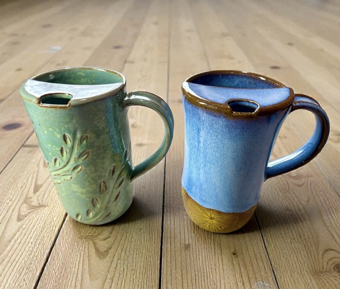 Two handmade ceramic mugs with spouts, one glazed in green with leaf patterns and the other in blue with decorative yellow design, placed on a wooden surface.