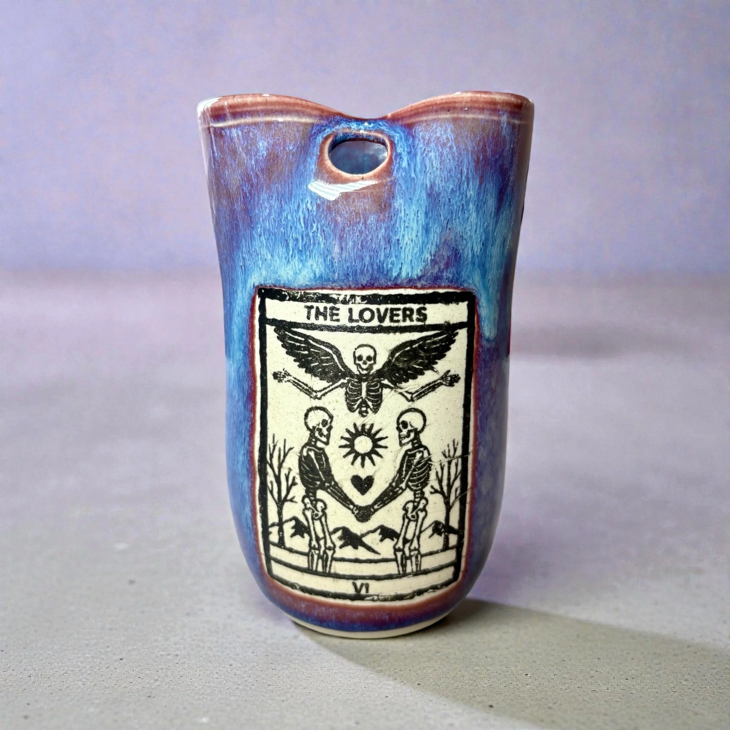 A ceramic vase with a blue and brown glaze, featuring a black and white illustration of skeletons and a skull with wings, titled "The Lovers".