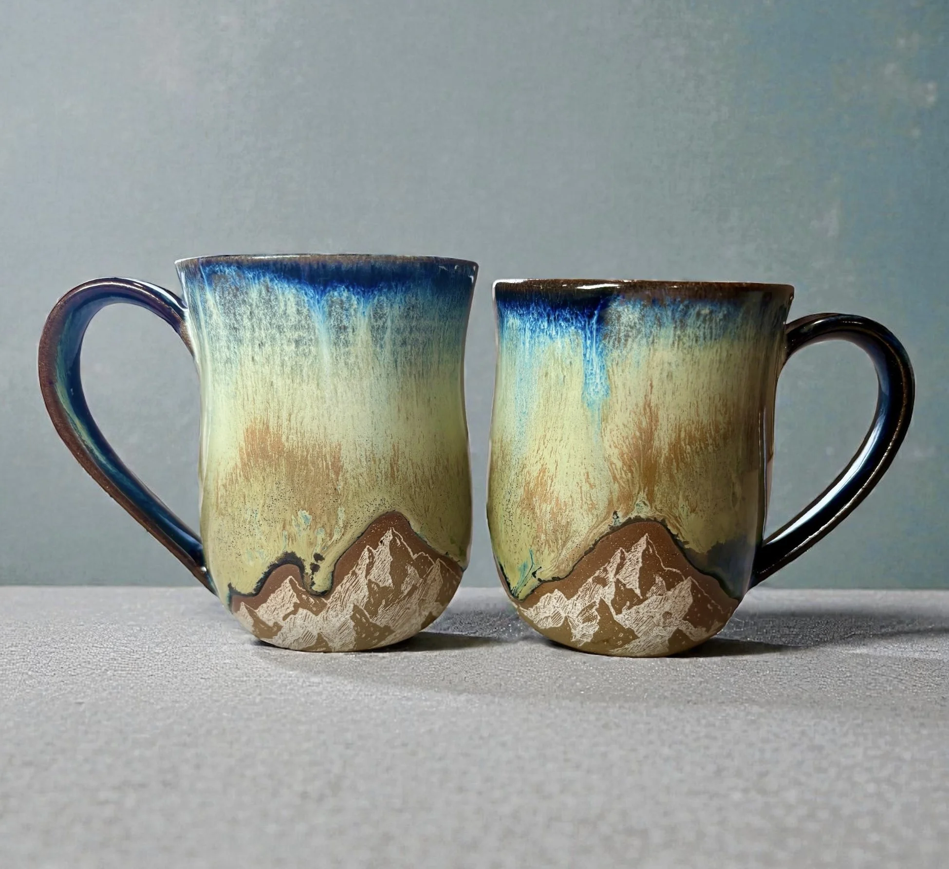 Two ceramic mugs with mountain and blue sky landscape paintings on them, placed on a light gray surface against a light blue background.