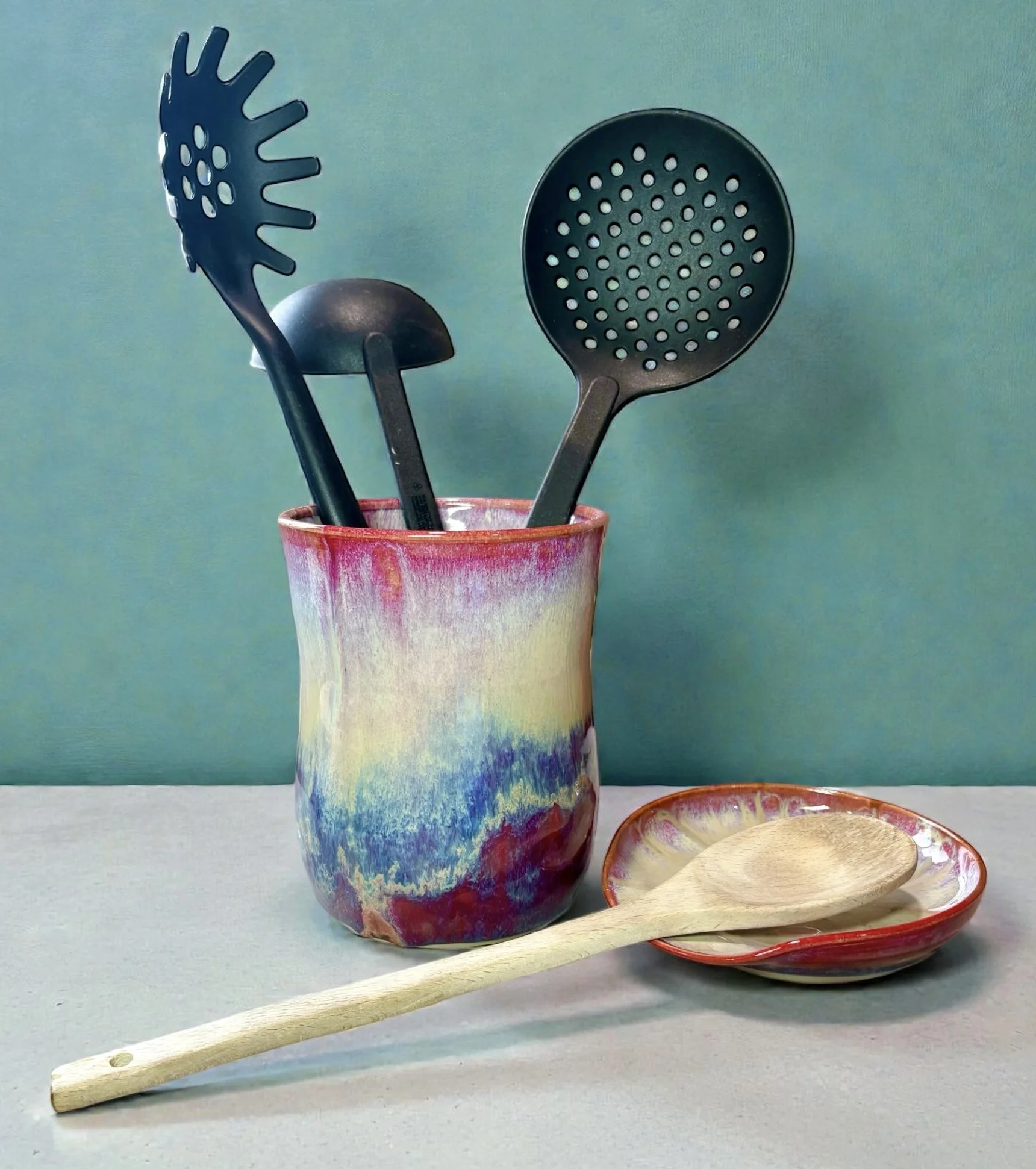 A colorful ceramic cup holding three black kitchen utensils: a slotted spoon, a ladle, and a slotted skimmer. A small ceramic dish and a wooden spoon are in the foreground on a white surface, with a plain teal background.