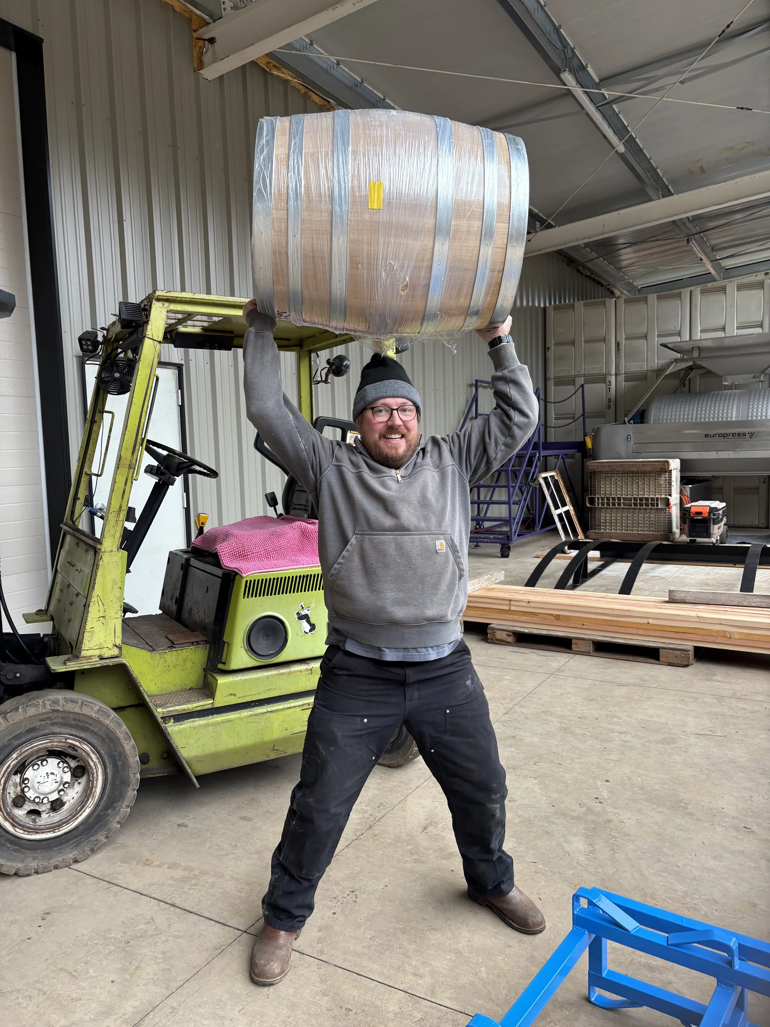 Man smiling and posing with a large wooden barrel on his shoulders inside a warehouse or industrial space. He is wearing glasses, a grey hoodie, black pants, and brown boots. There is a yellow forklift behind him, along with various items including a pink towel, crates, and tools.