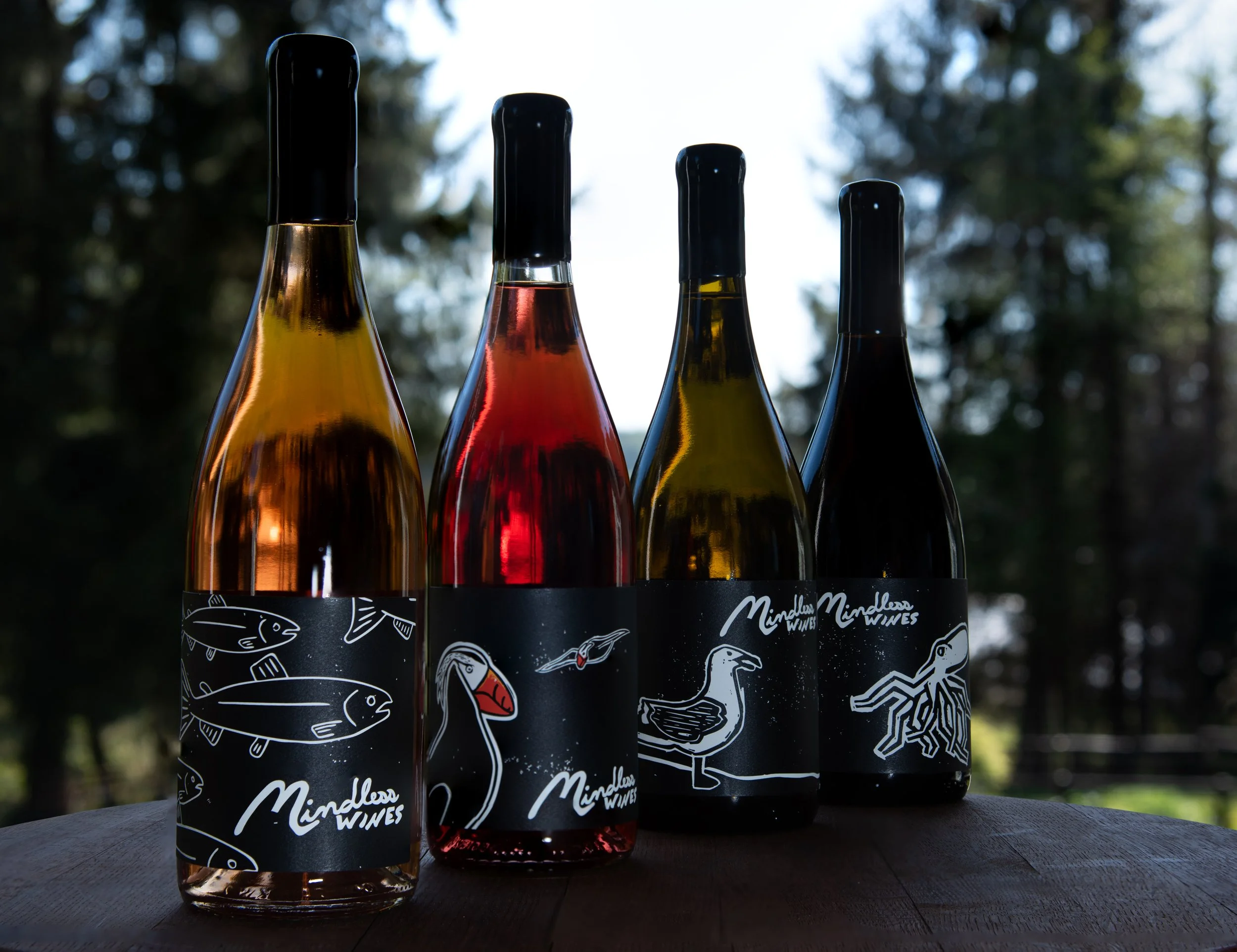 Four wine bottles from the willamette valley with colorful liquids, decorated with illustrated fish, bird, and crustacean designs on black labels, placed outdoors on a dark wooden surface with trees in the background.