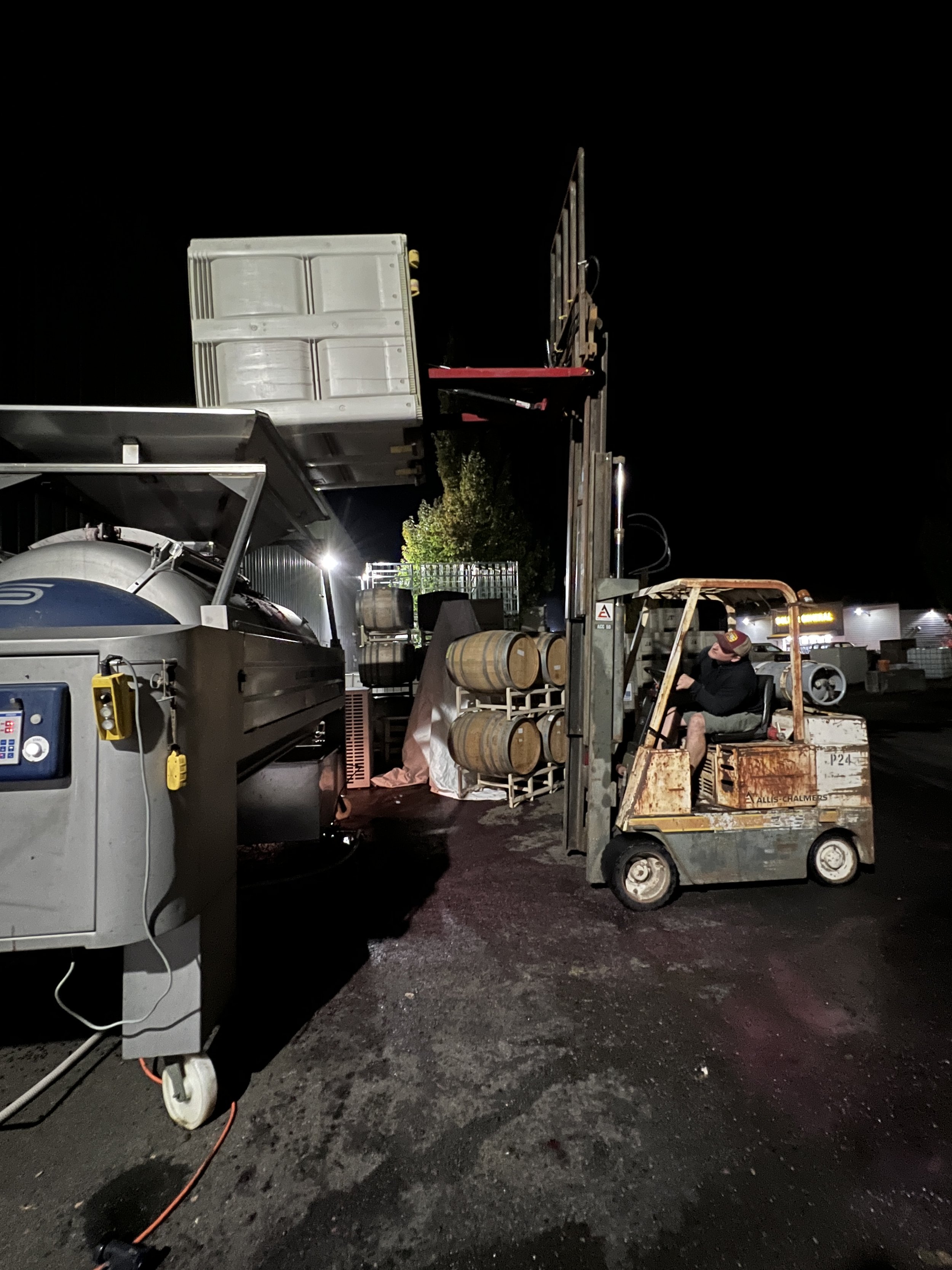 Night scene in a parking lot with a worker operating a small forklift, barrels and equipment around, illuminated by bright lights. dumping grapes into a wine press