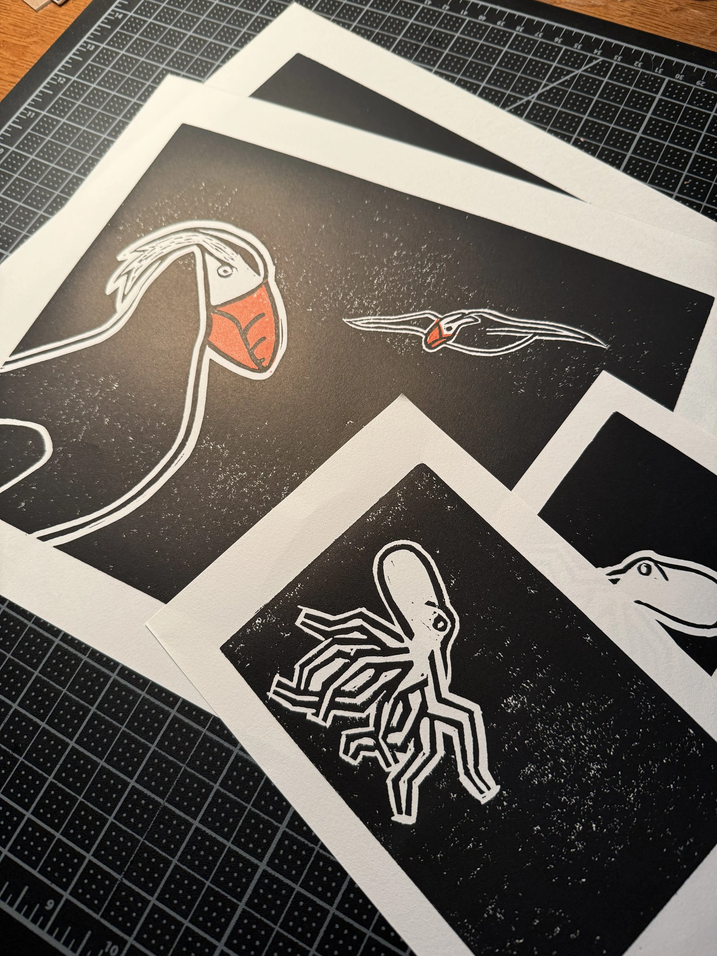 Printed block prints of a Puffin, a bird in flight, and a octopus on white paper sheets, placed on a black cutting mat with grid lines.