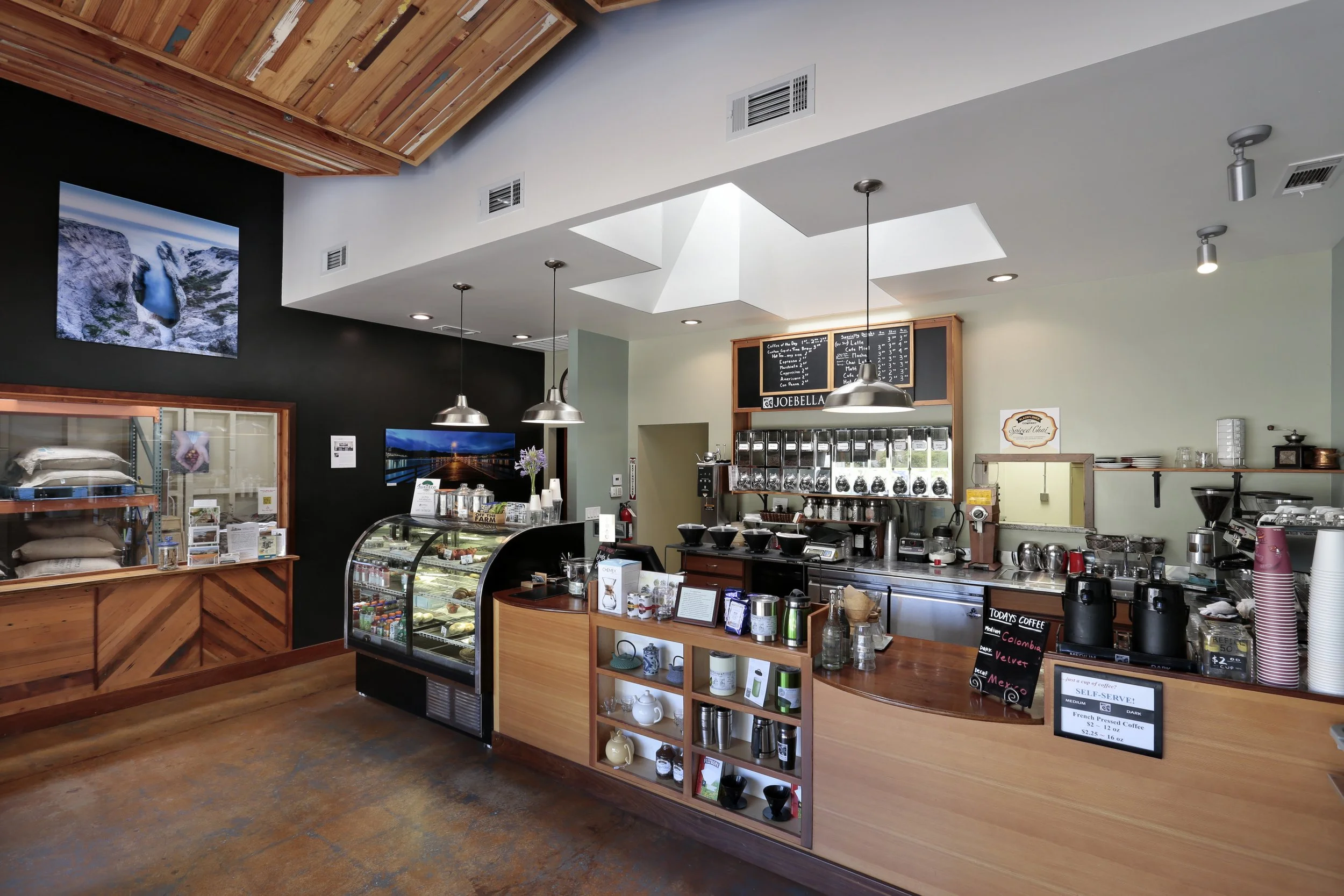 Interior of a cozy coffee shop featuring a wooden counter, espresso machines, a menu board, and decor with paintings of landscapes, warm lighting, and a display case of desserts.
