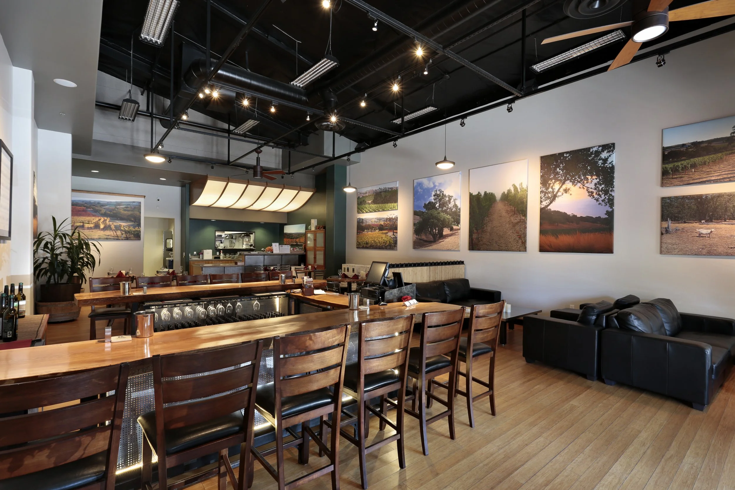 Interior of a modern restaurant or cafe with a bar counter, wooden chairs, black leather couches, and landscape photographs on the wall.