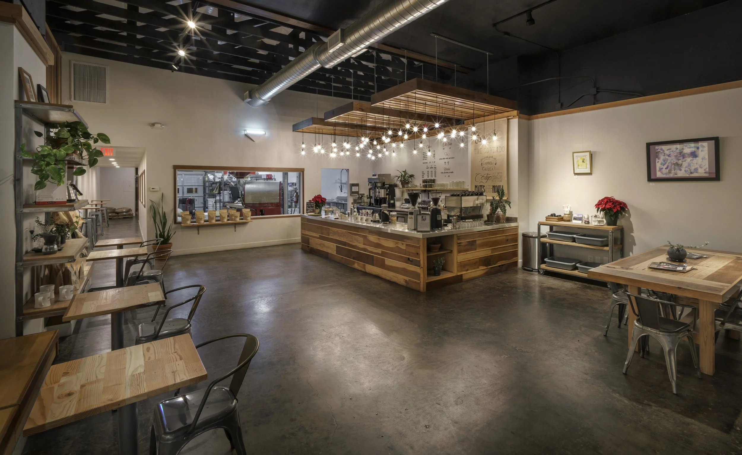Interior of a modern cafe with wooden tables, chairs, and a coffee counter. Bright lighting, potted plants, and artwork on the walls create a cozy atmosphere.
