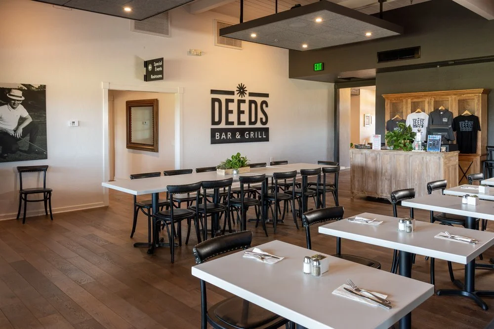 Interior of Deeds Bar and Grill with tables, chairs, and a wooden bar counter, featuring a sign and merchandise display.
