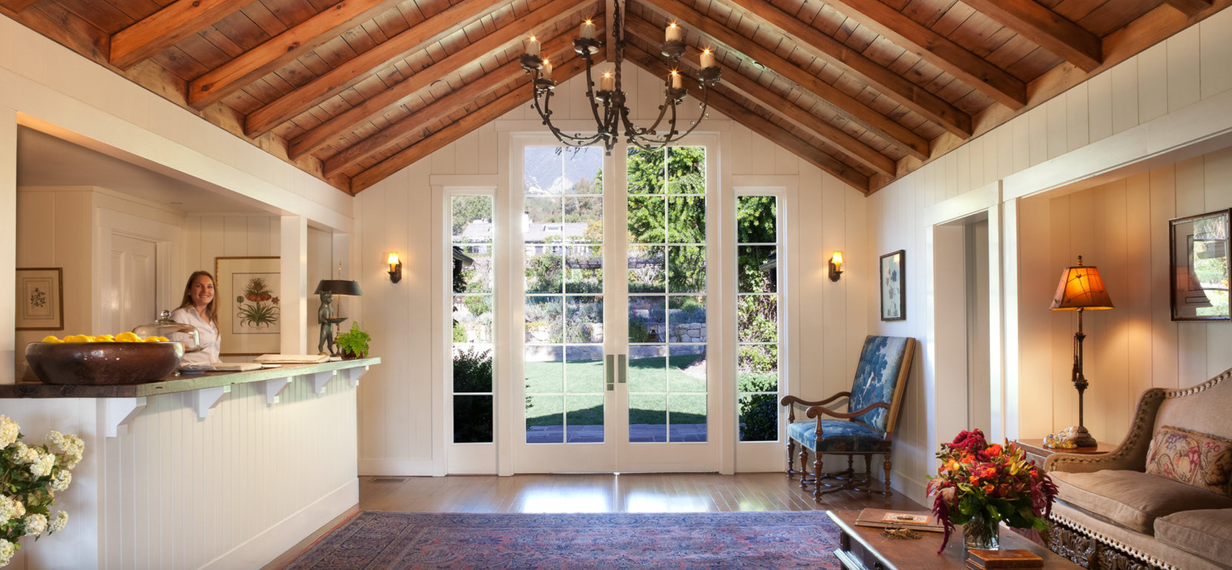 Living room with a high wooden ceiling, large windows, and outdoor view. A woman stands behind a white kitchen counter smiling. There is a blue patterned armchair, a floral sofa, and a table with a vase of flowers. Warm lighting and decor create a co