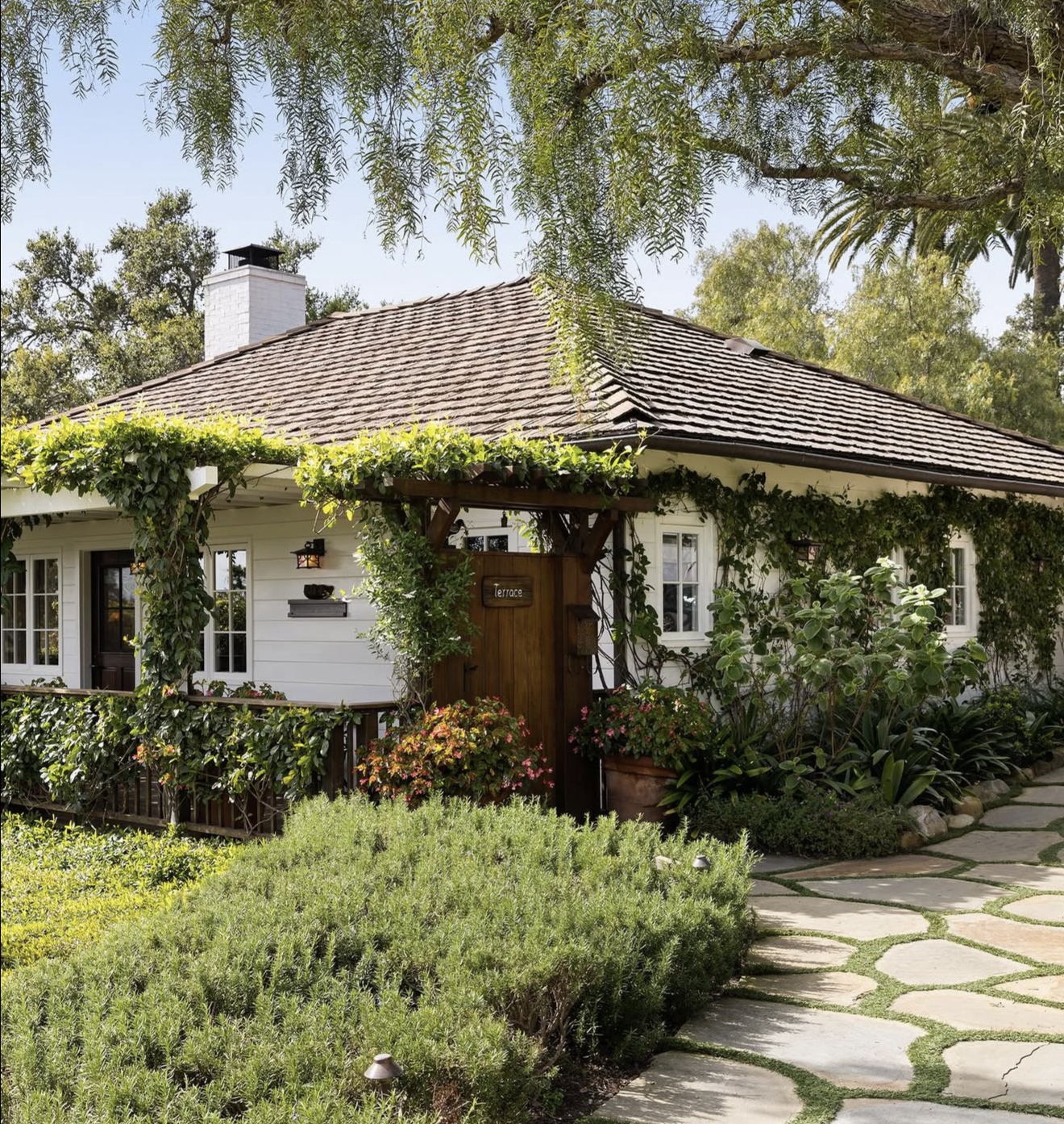 A cozy white house with a shingle roof, surrounded by lush greenery, plants, and a stone pathway.