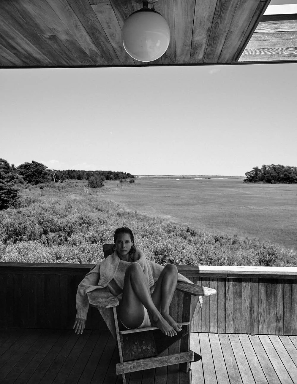 A woman sitting in a wooden chair on a porch overlooking a wide open landscape with trees and grass, under a wooden ceiling with a hanging light fixture.