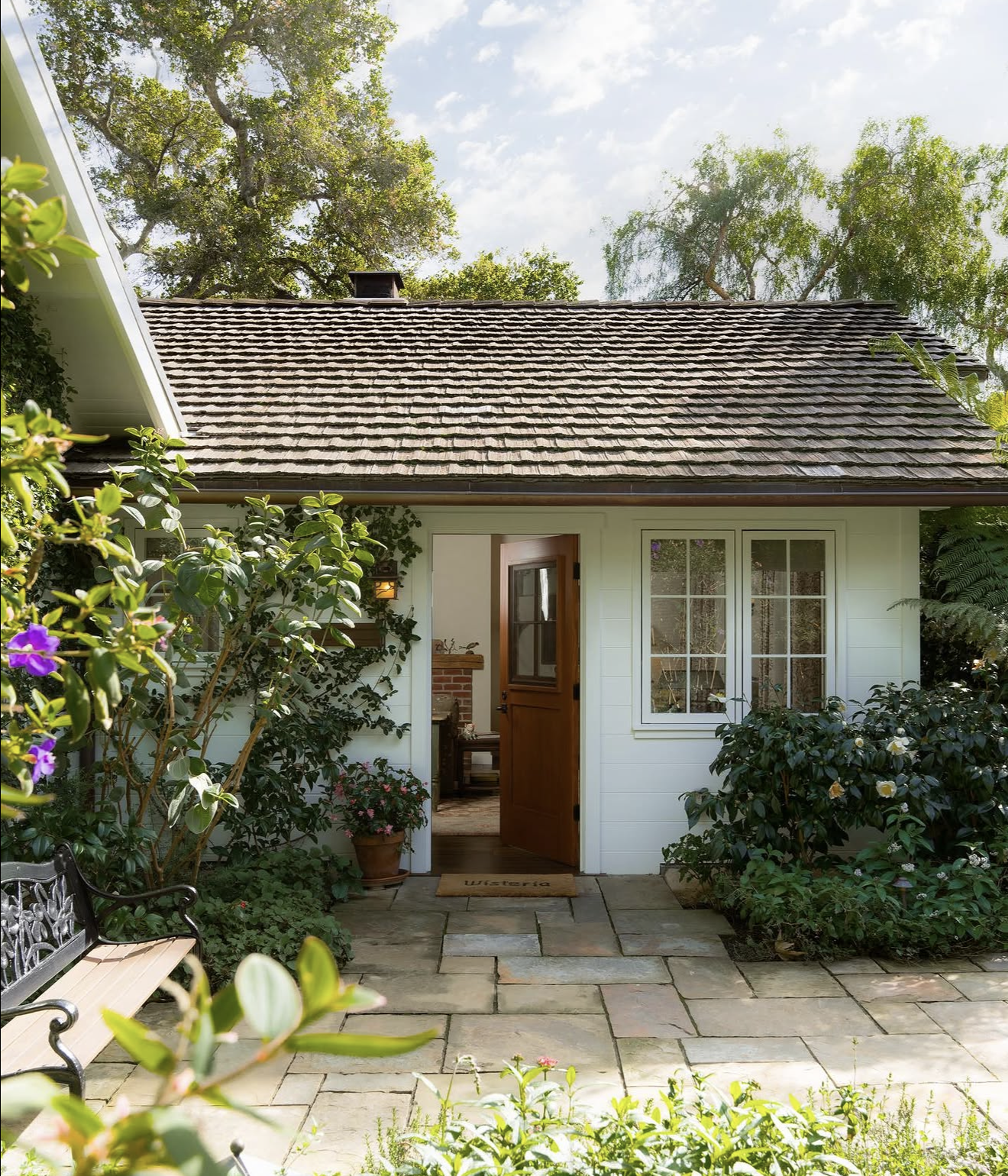 Front view of a cozy white cottage with a patio, open wooden door, and lush garden with green shrubs and patio stones. Large trees are in the background under a partly cloudy sky.