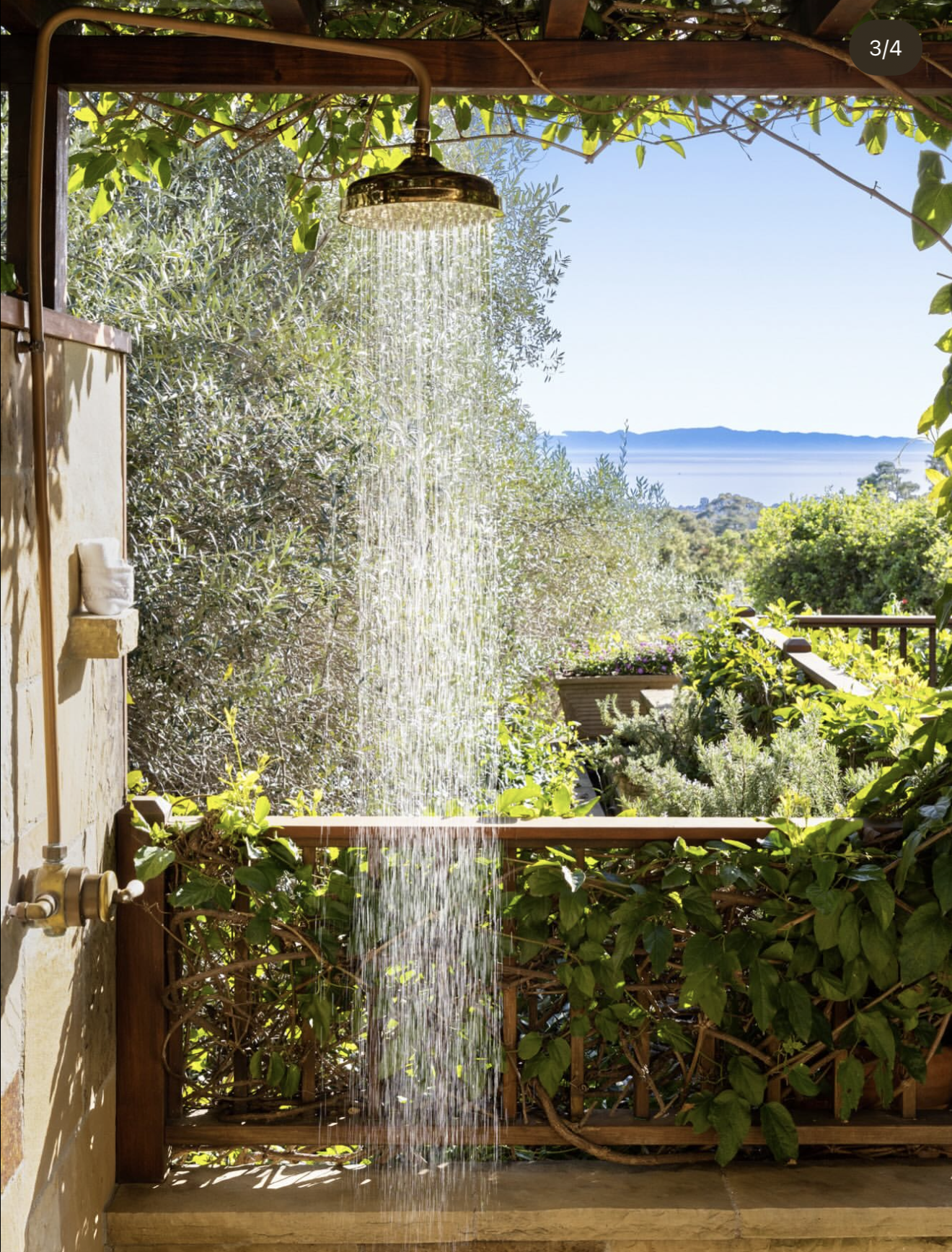 Outdoor shower with a waterfall feature surrounded by greenery, overlooking a scenic landscape with water and mountains in the distance.