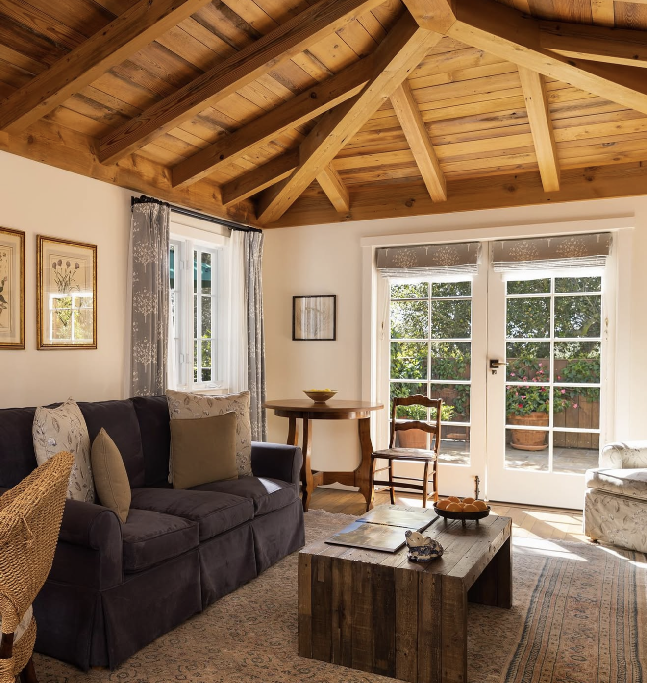 Living room with a vaulted wooden ceiling, a dark couch with decorative pillows, a wooden table, and large window and glass doors showing an outdoor patio with potted plants.