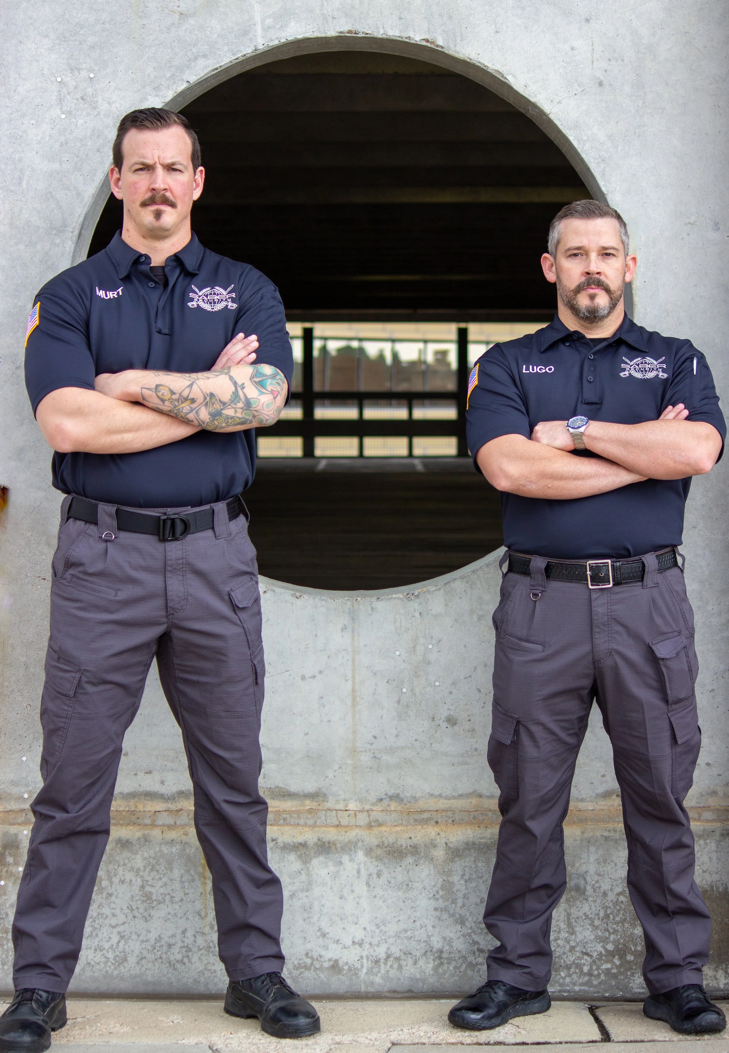 Two security personnel standing confidently with crossed arms in front of concrete structure with large arched opening.