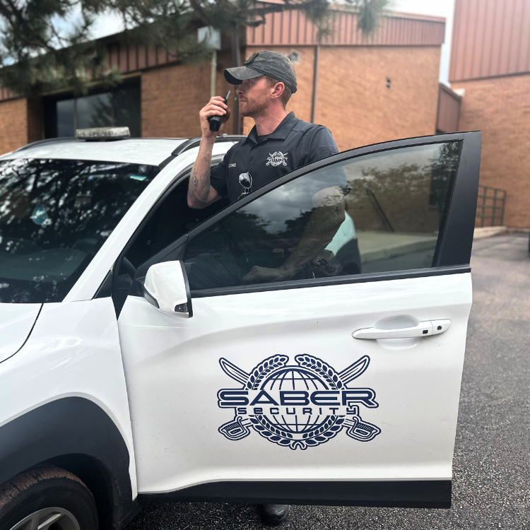 A security officer stands next to a white security vehicle with the SABER Security logo on the door, talking on a walkie-talkie.