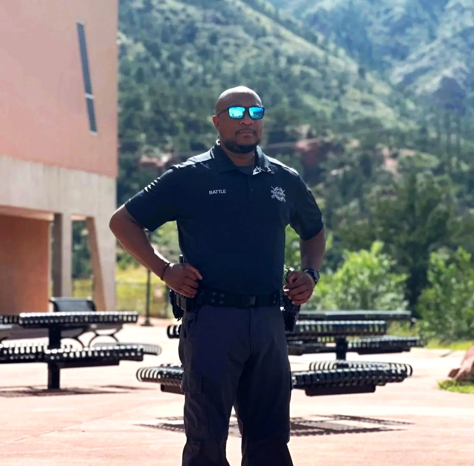 A police officer standing outdoors with sunglasses, with a scenic backdrop of green hills, trees, and a building to the left. The officer has hands on his hips and is wearing a Saber Security uniform.