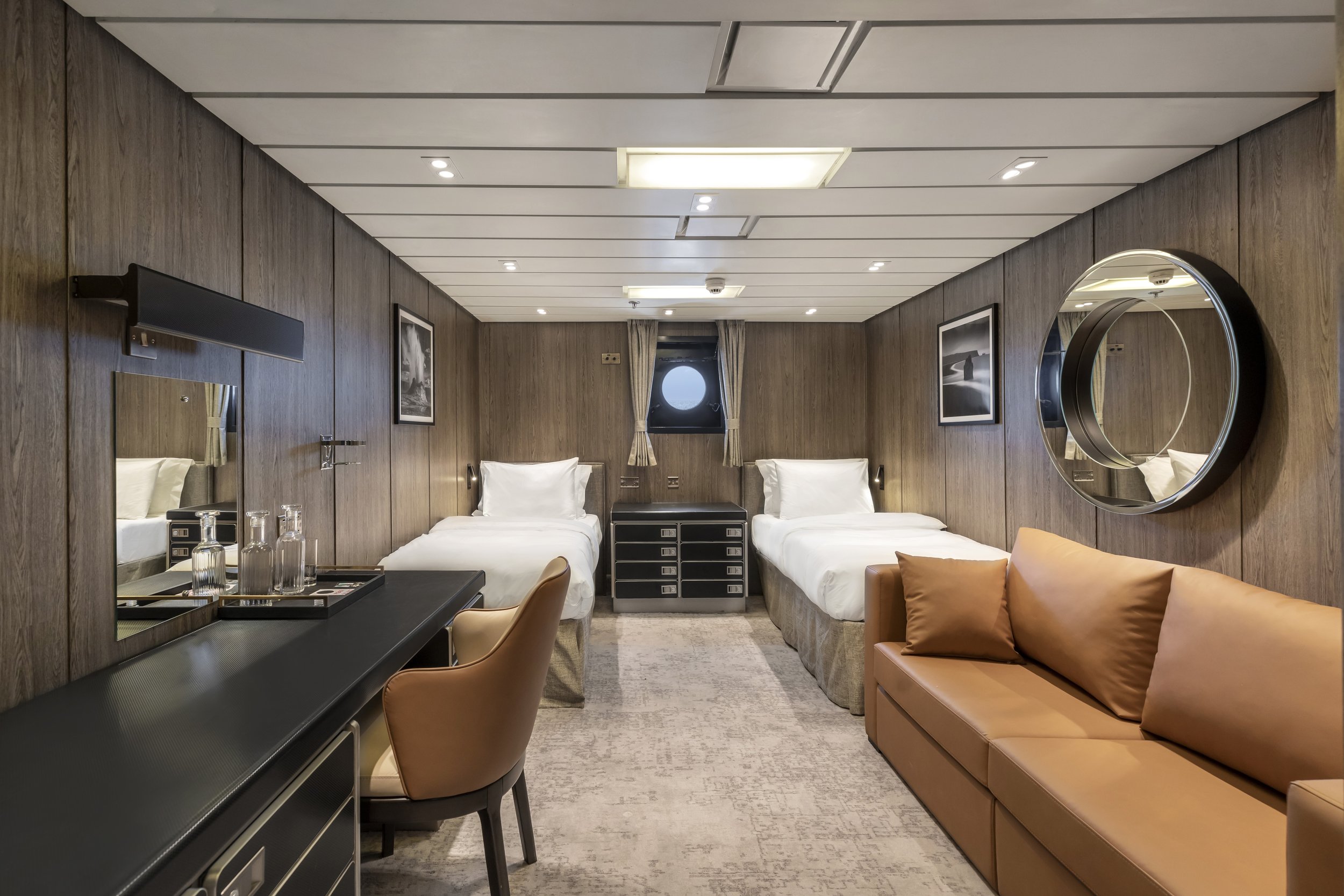 A modern cruise ship cabin with two single beds, a brown sofa, and a wooden wall with artwork and mirrors.
