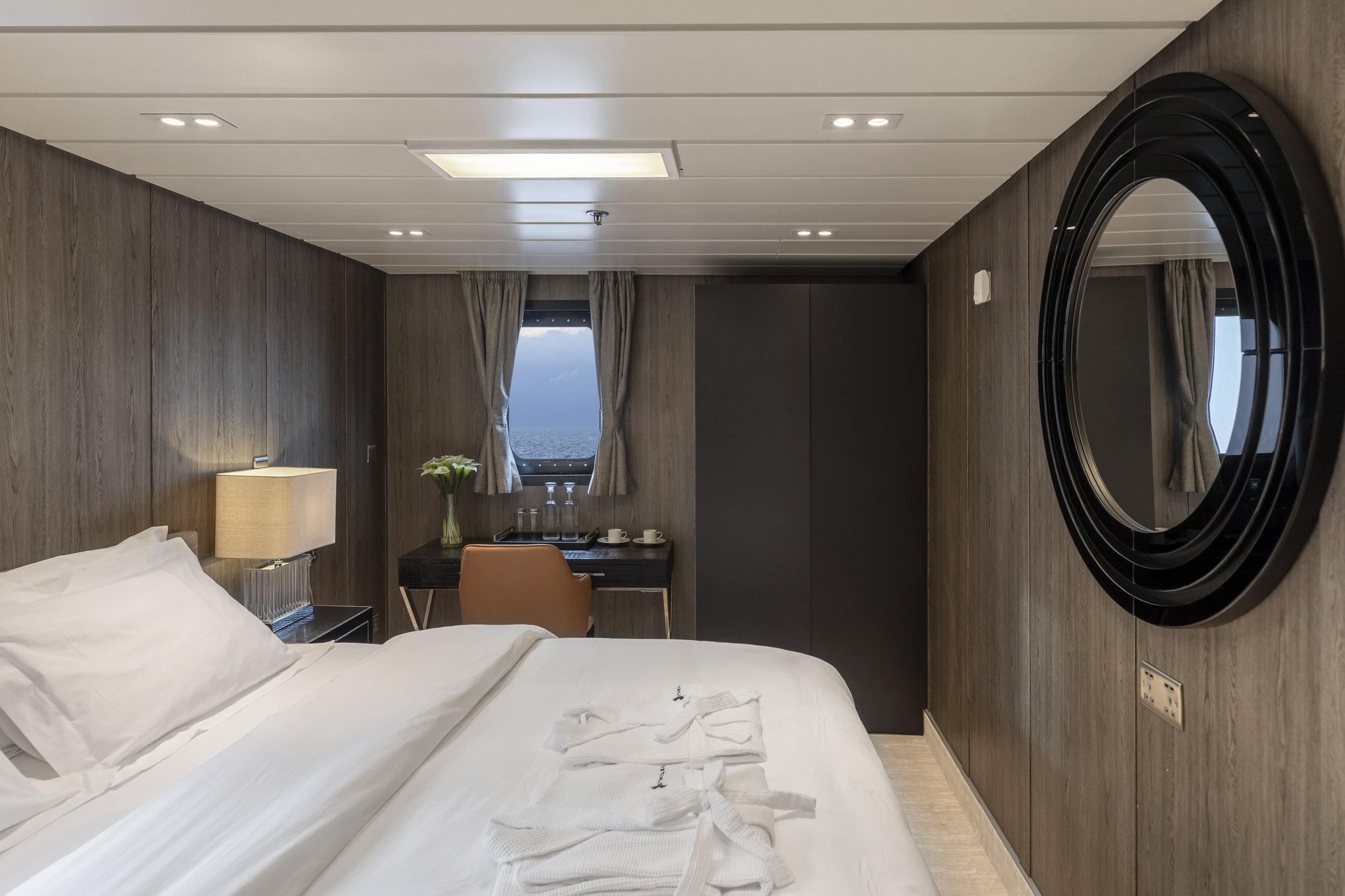 Interior of a hotel room featuring a bed with white linens, a nightstand with a lamp, a small desk with a chair, a window showing the ocean outside, a large round mirror on the wall, and various small decorative items.
