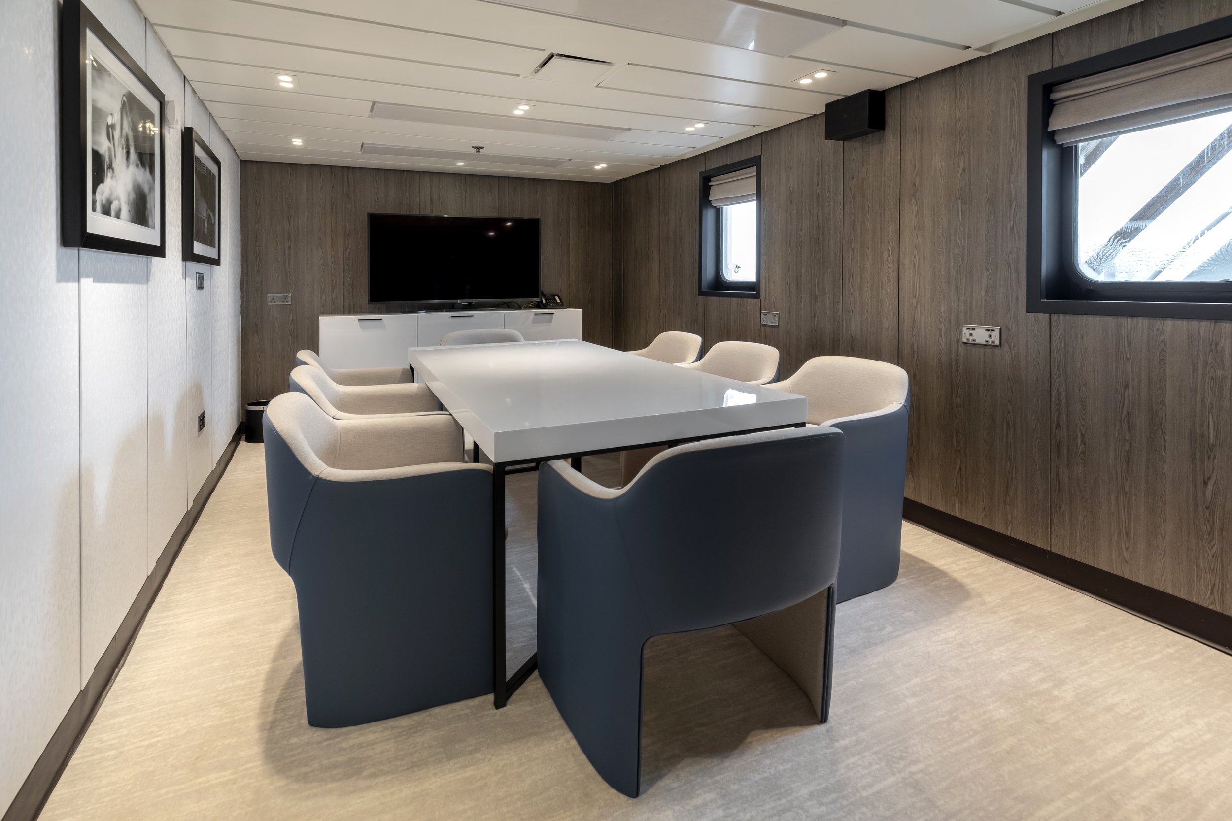 Modern conference room with eight beige and navy blue chairs around a rectangular white table, wall-mounted black flat-screen TV, wooden and white wall panels, and two windows with blinds.