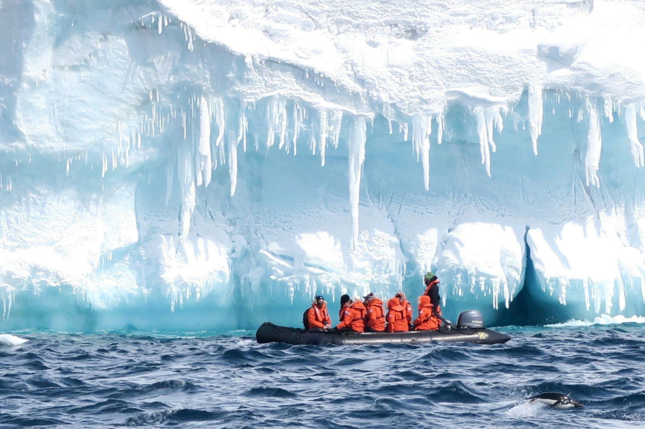 Group of people in orange life jackets on a black inflatable boat near an iceberg with icicles hanging down.