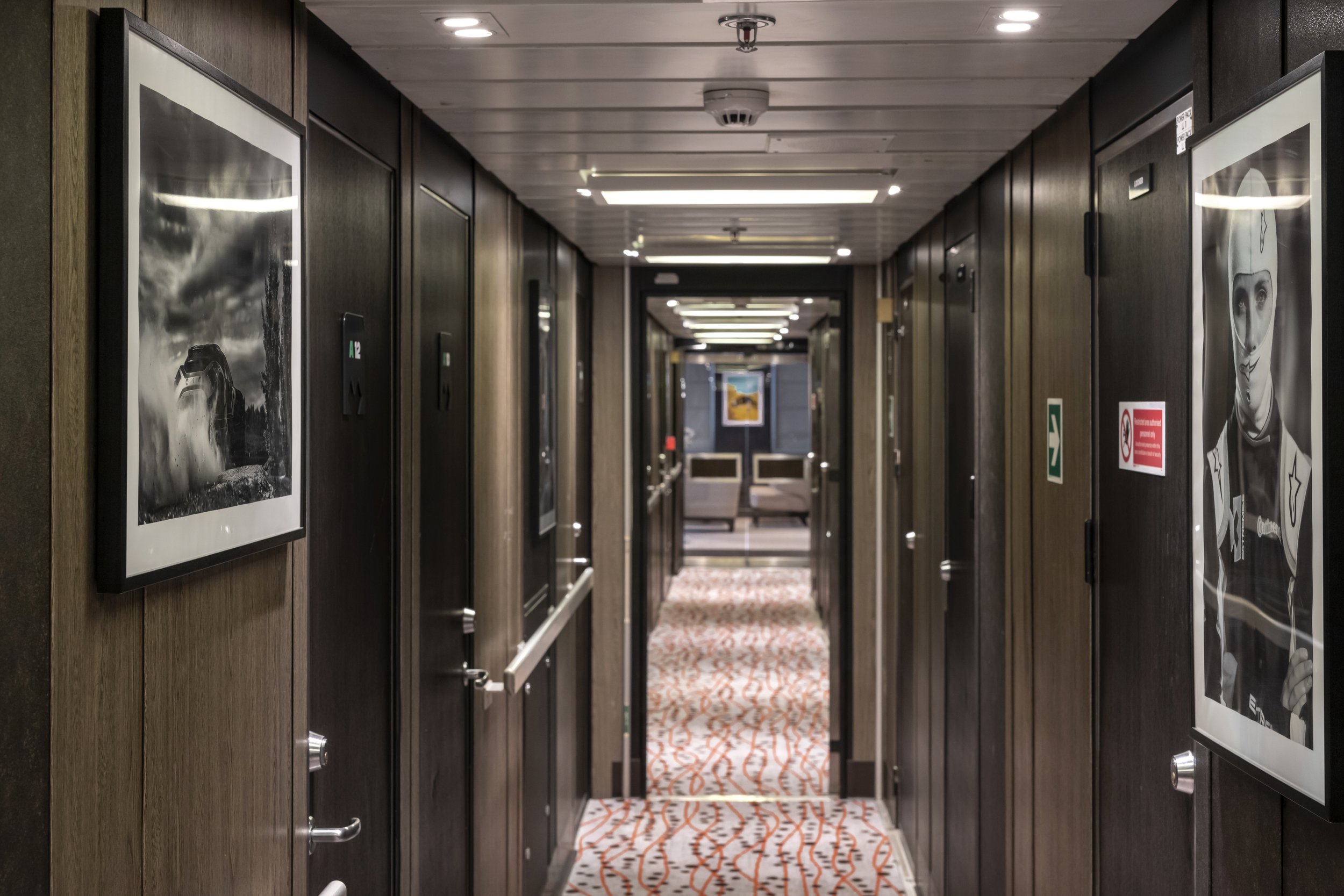 Long hotel corridor with dark wood walls, framed artwork, patterned carpet, and seating area at the far end.