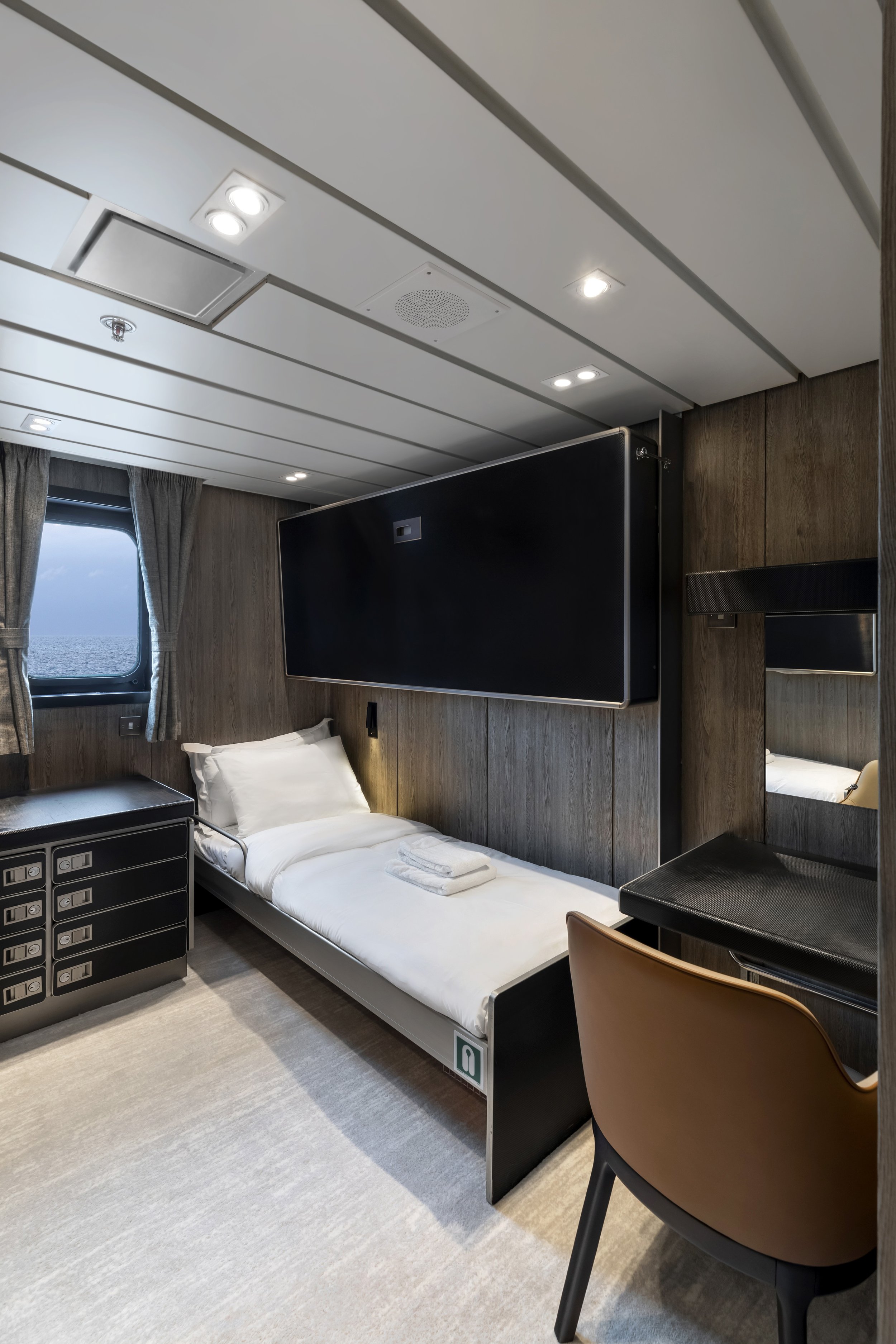 A modern cruise ship cabin with a small bed, a window showing the ocean, a black dresser, a mirror, a chair, and a wall-mounted television.